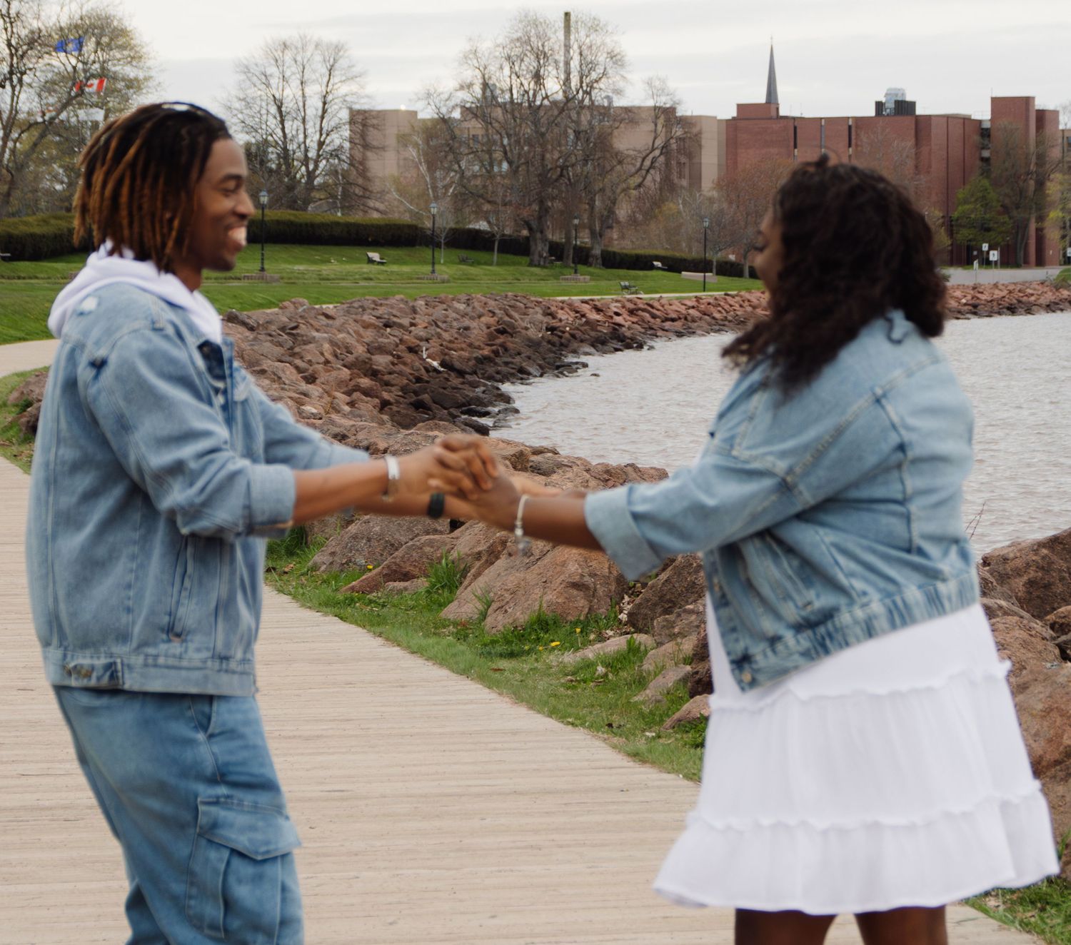 Two people in denim jackets dance joyfully by the waterfront.