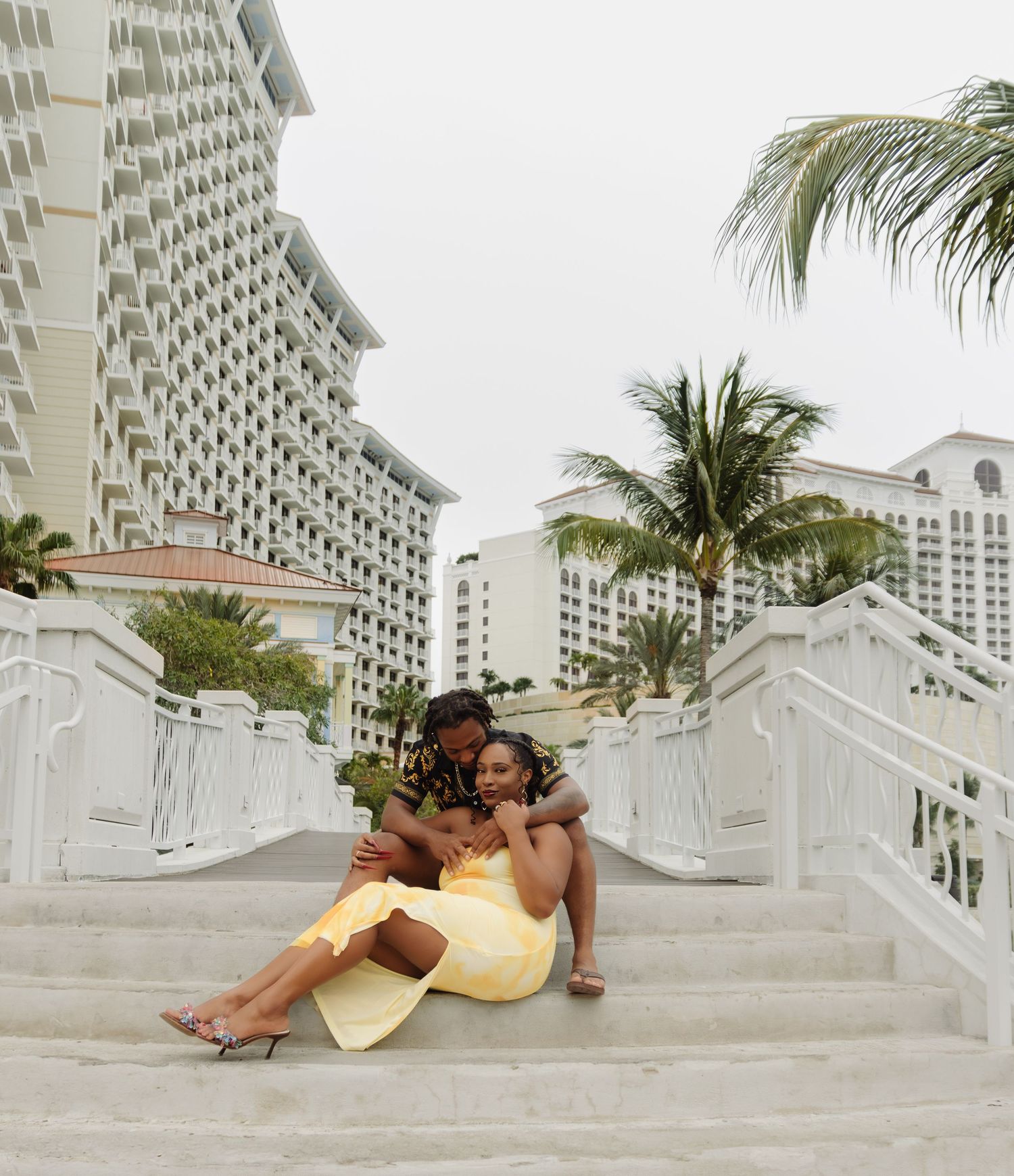 A couple relaxes on white steps with palm trees and resort buildings in the background.