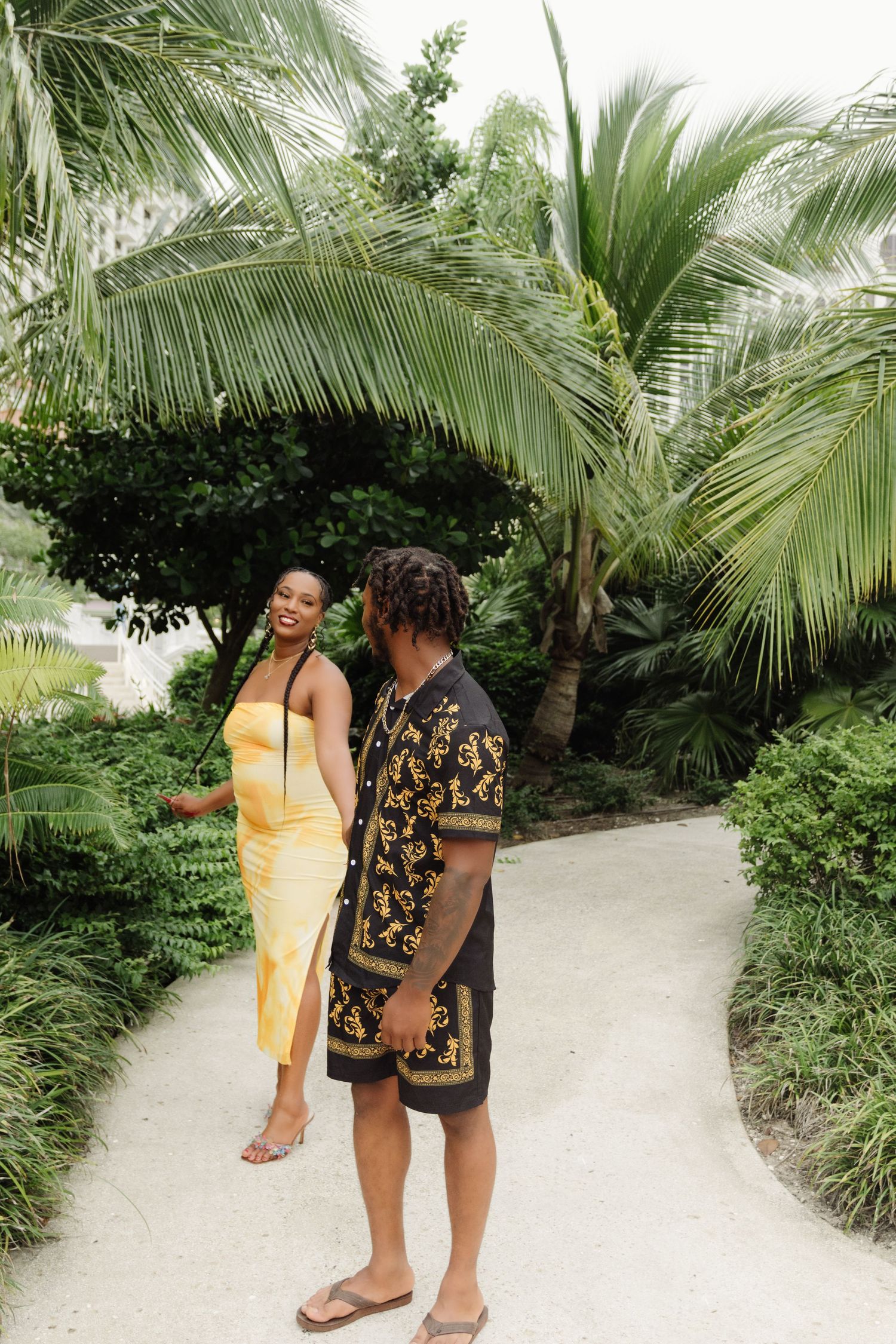 A stylish couple walking along a palm-lined pathway in coordinated outfits.