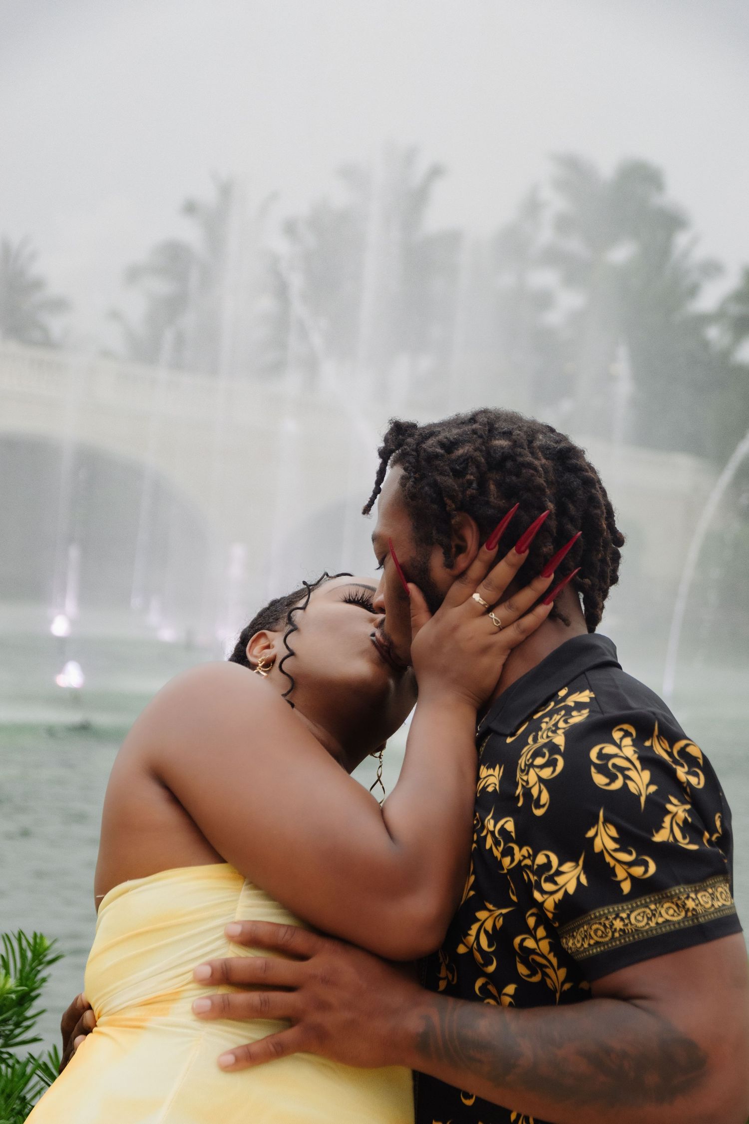 A romantic couple shares a kiss near a fountain in a misty garden setting.