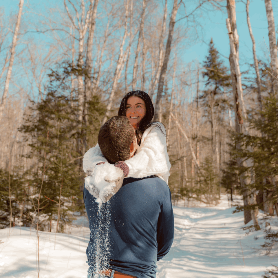 A romantic winter scene shows a couple embracing on a snowy trail surrounded by bare birch trees and evergreens.