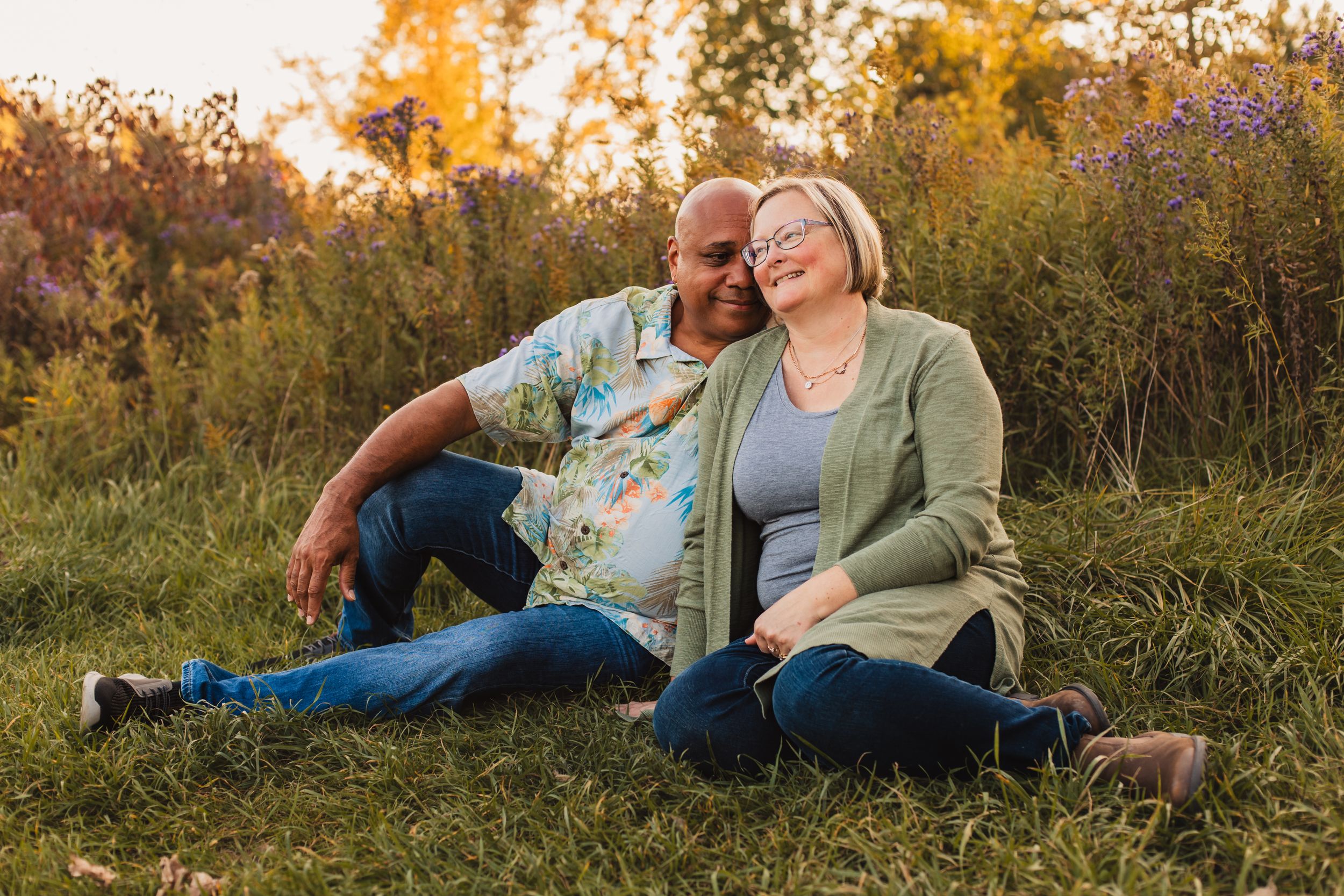 A couple sits together on grassy ground during golden hour sunset with autumn foliage in the background.