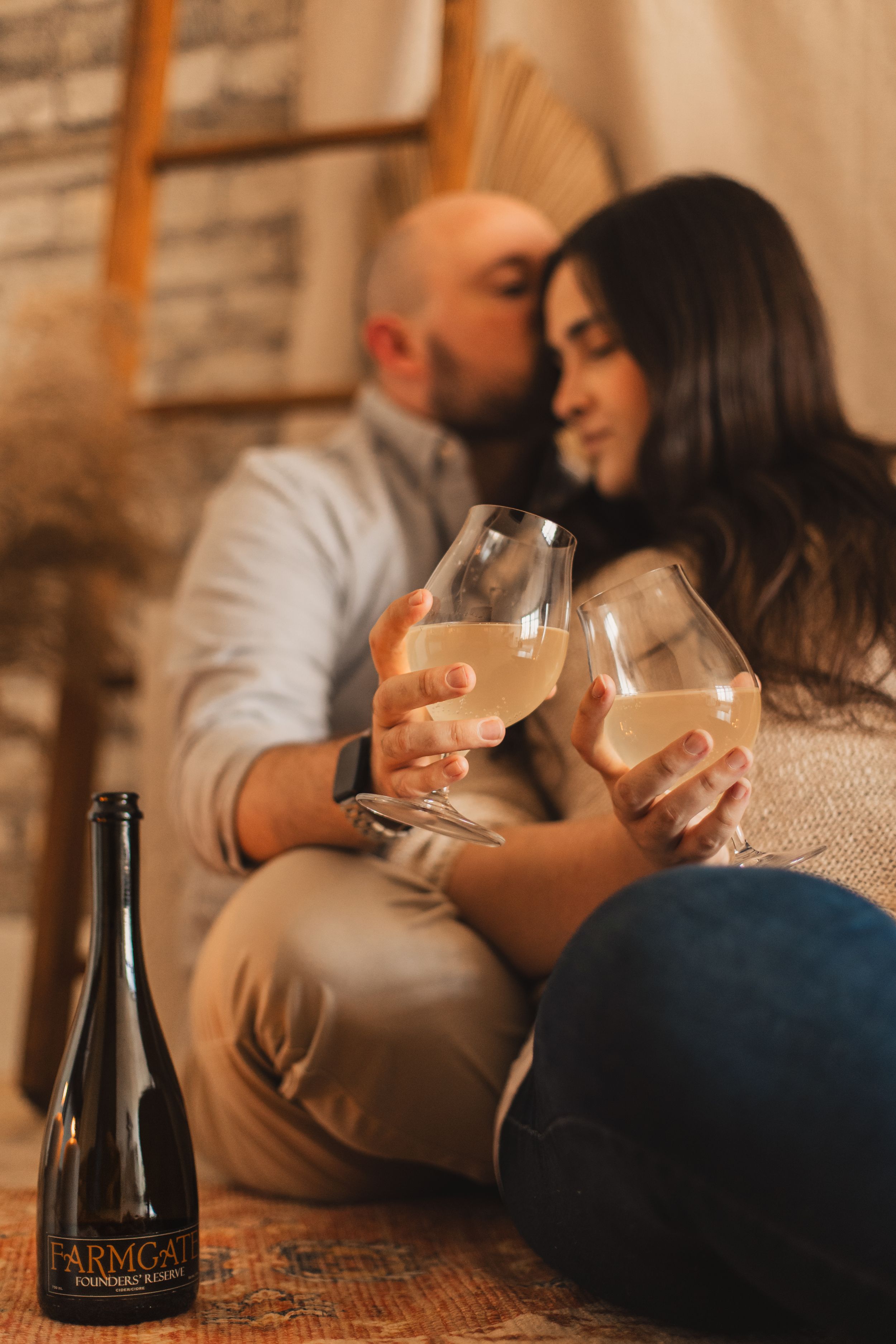 A couple relaxes on the floor with wine glasses while a dark bottle sits in the foreground.
