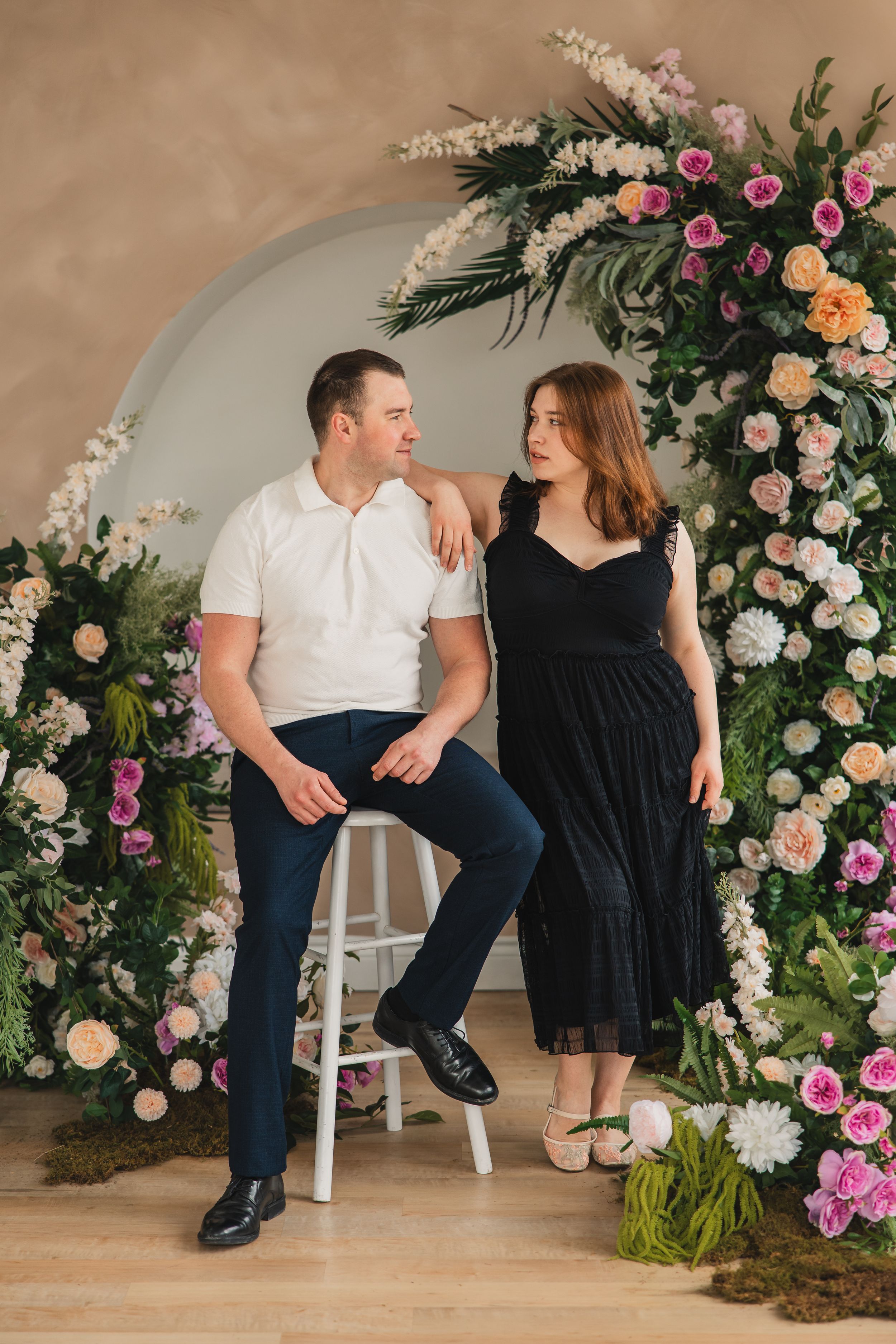 A romantic couple sits together on white stools surrounded by an elegant floral arch with pink and peach roses.