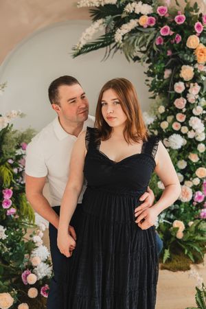 A couple poses together against a floral backdrop with pink and white roses and greenery in a romantic setting.