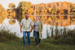 A couple walks along a serene lakeside path during golden hour as autumn colors reflect on the water's surface.
