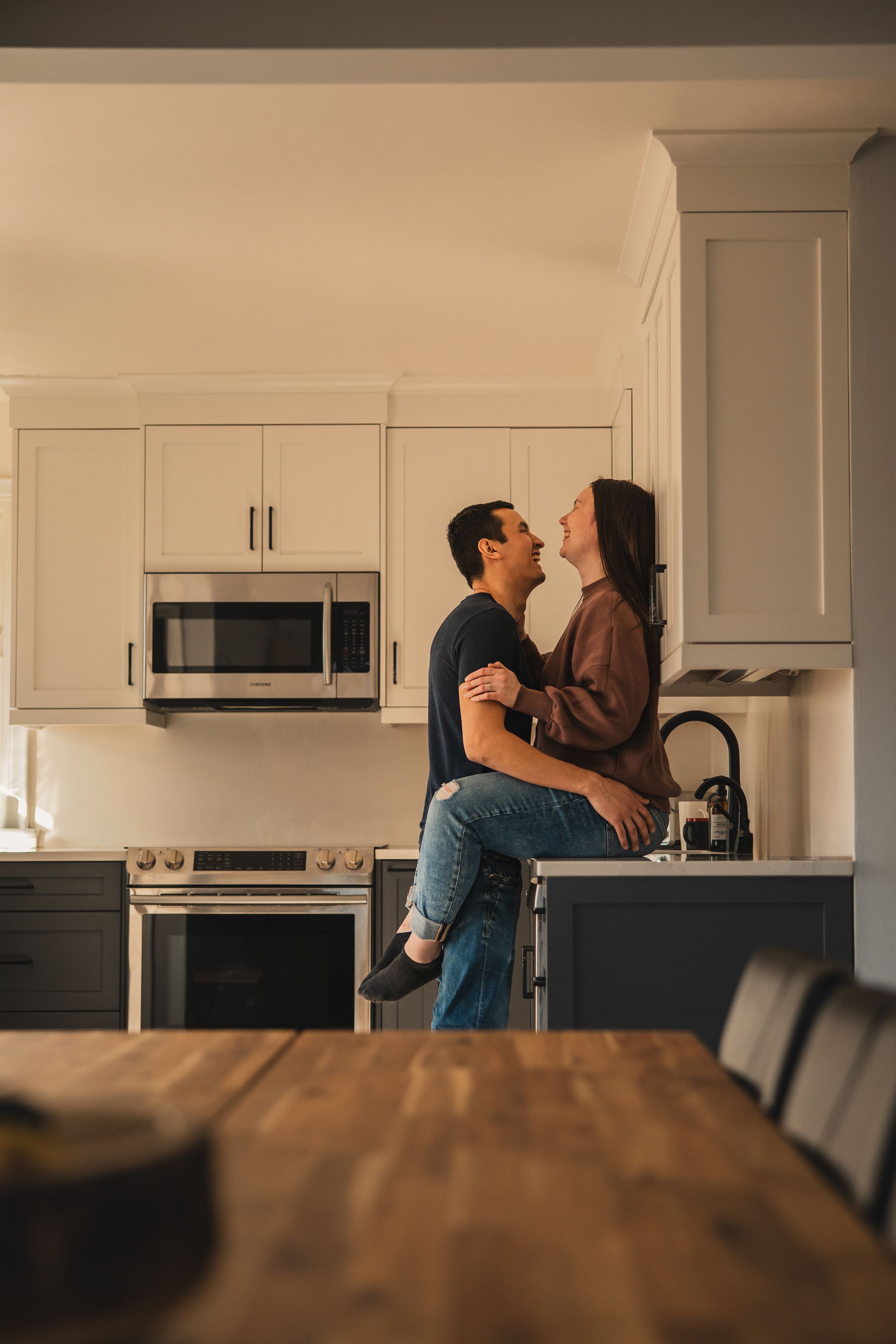 A couple shares an intimate moment dancing together in their modern white kitchen with black appliances.