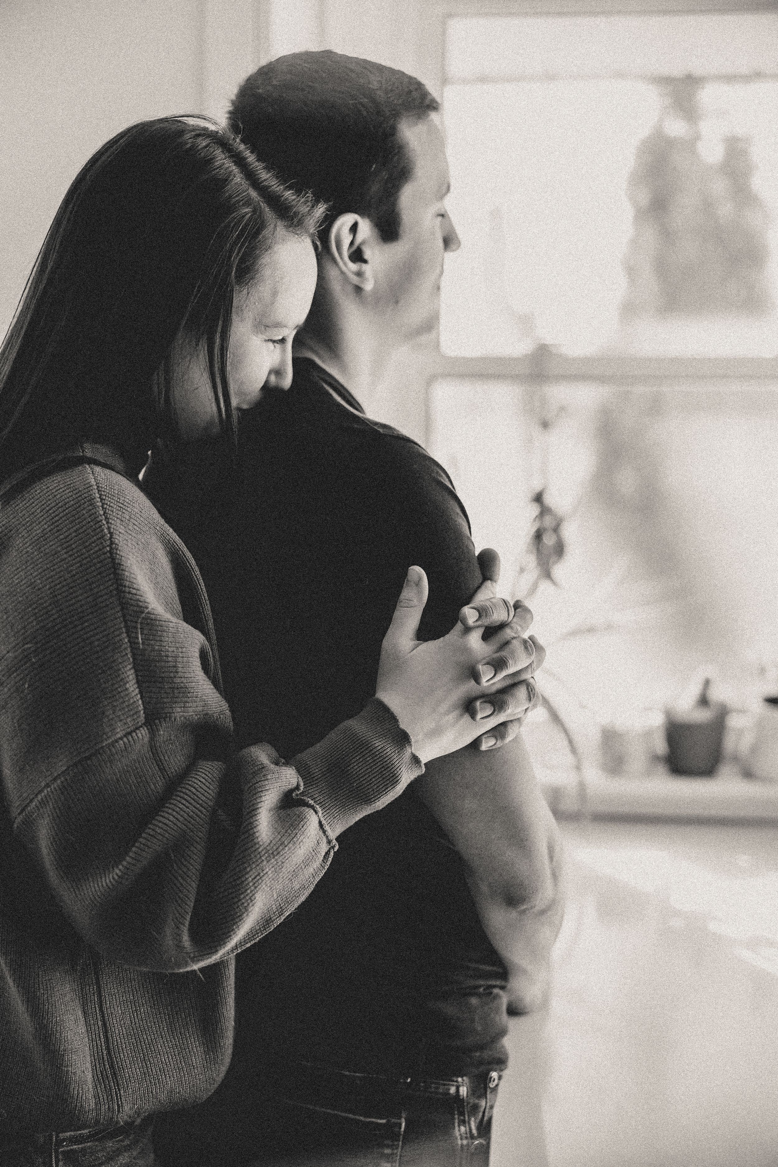 A romantic black and white photo of a couple embracing by a window, with the woman hugging from behind.