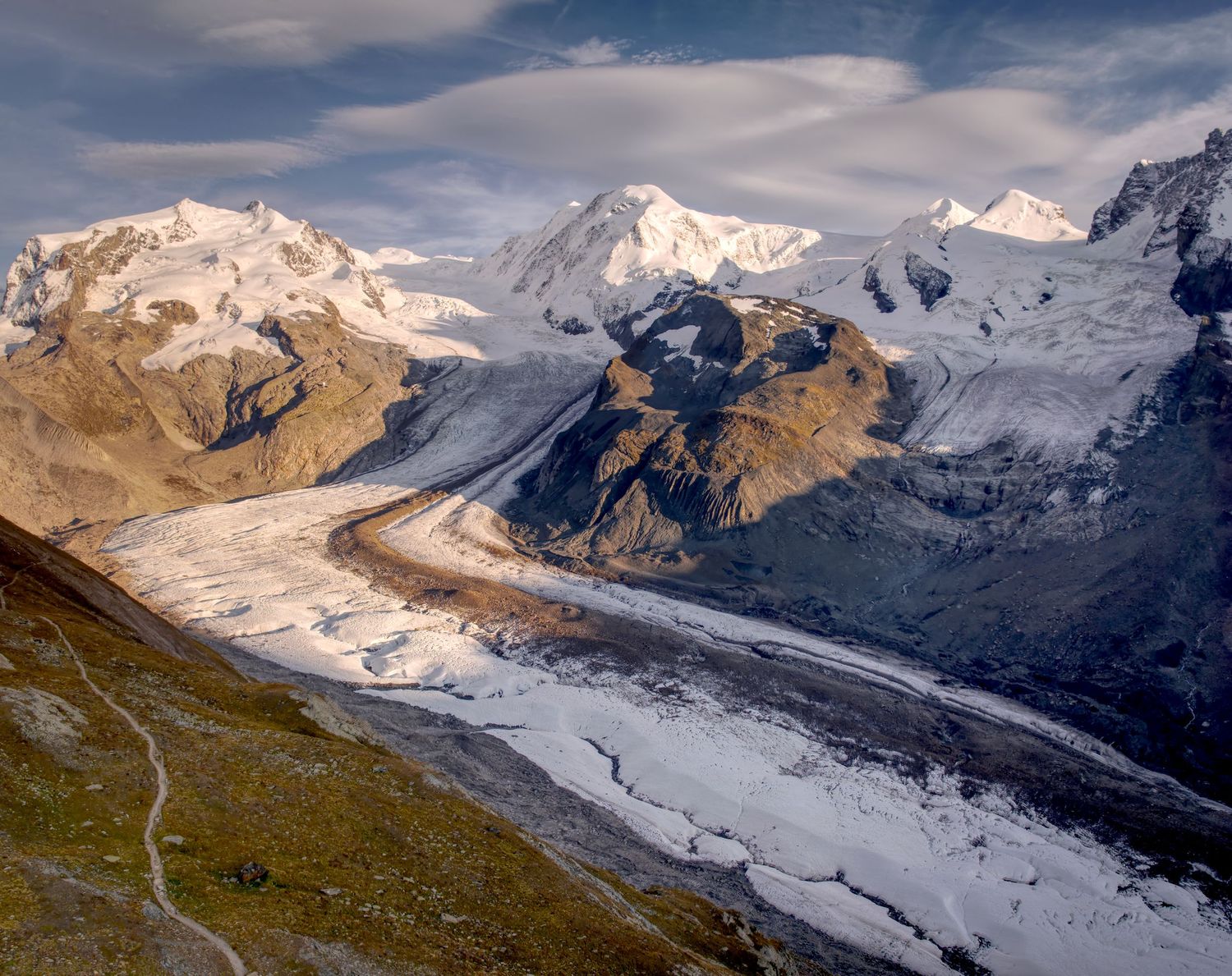 Towering glacier mountain peaks with sun and clouds over glacier stream.