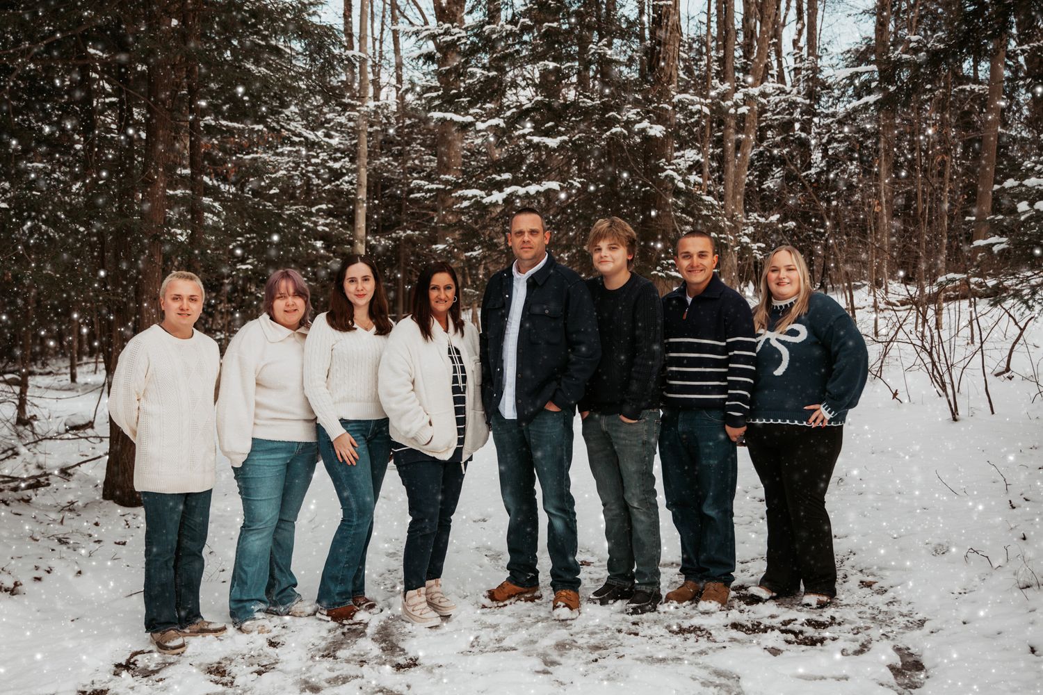 A group of eight people pose together in a snowy wooded area wearing coordinated white and dark clothing.