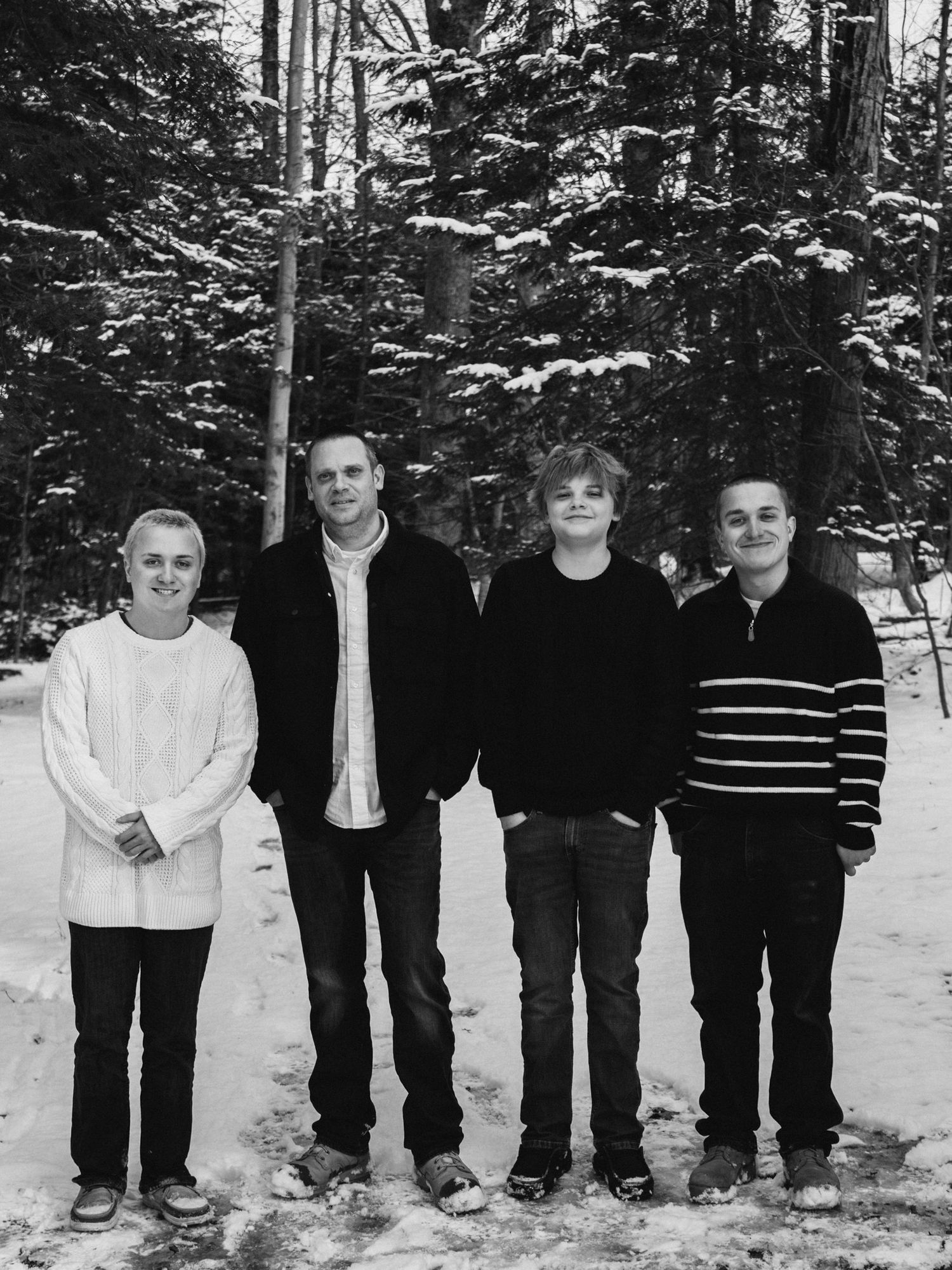 Four people stand together in a snowy forest setting wearing casual winter clothing.