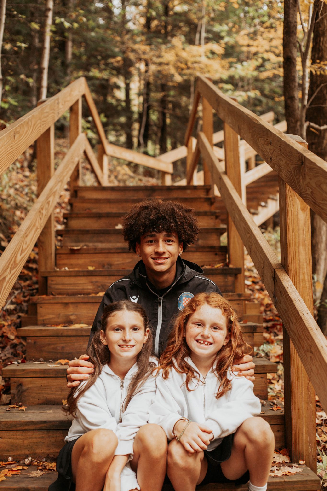 Three children in white shirts sit together on wooden steps surrounded by autumn foliage.