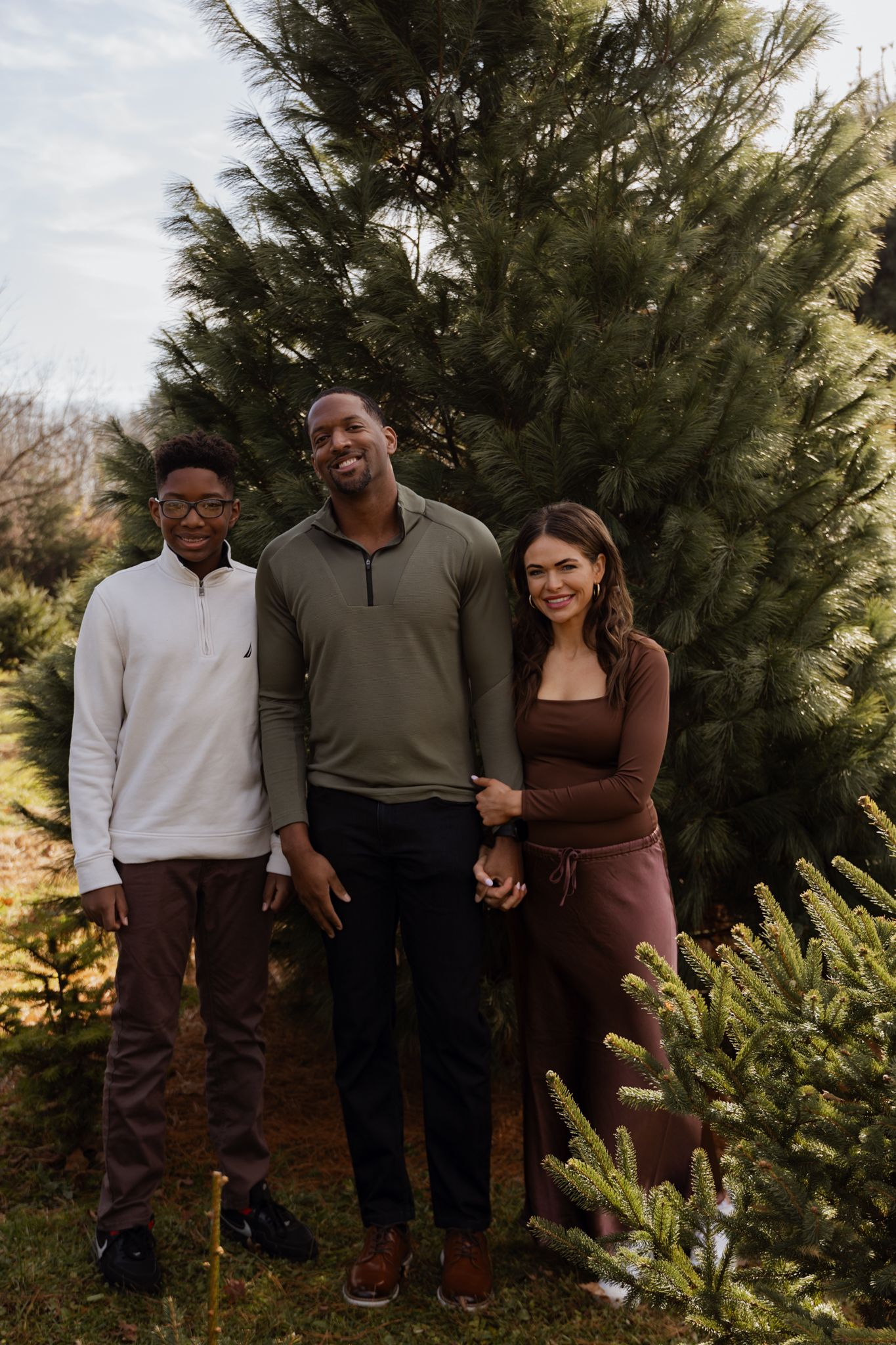 Three people pose together in front of evergreen trees during a winter outdoor photoshoot.