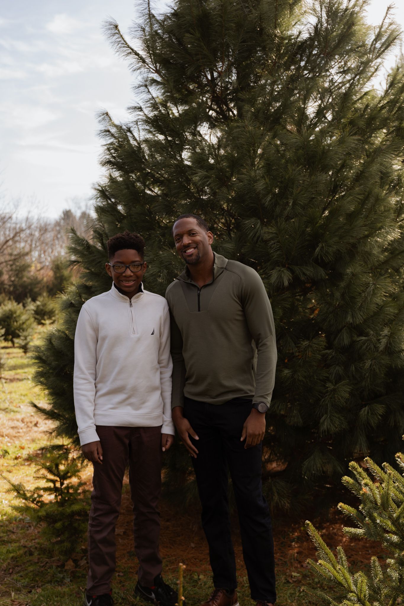 Two people stand together smiling in front of evergreen trees at a Christmas tree farm on a sunny autumn day.