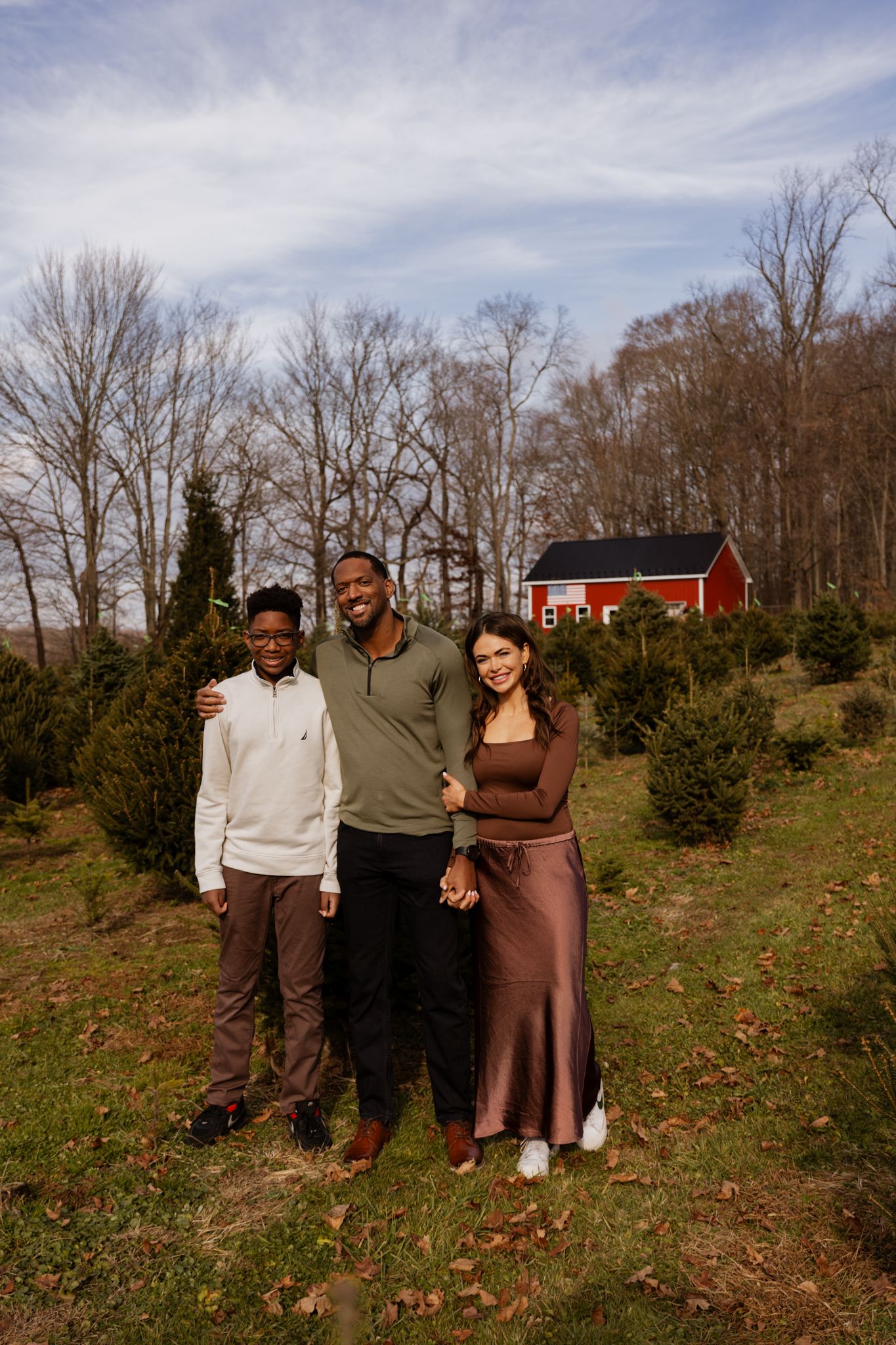 Three people stand together in a rustic outdoor setting with bare trees and fallen leaves in autumn.