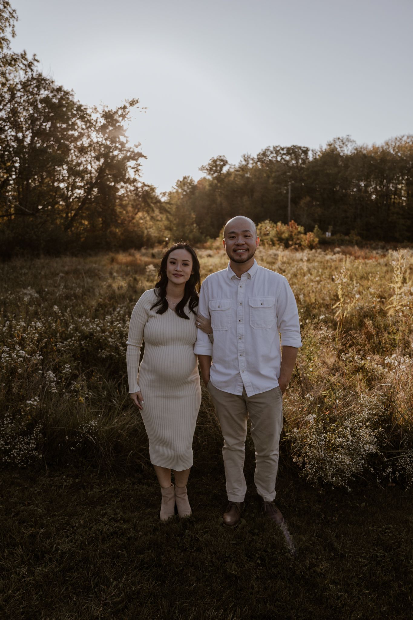An expecting couple poses together in a golden sunset field wearing coordinated light colored outfits.