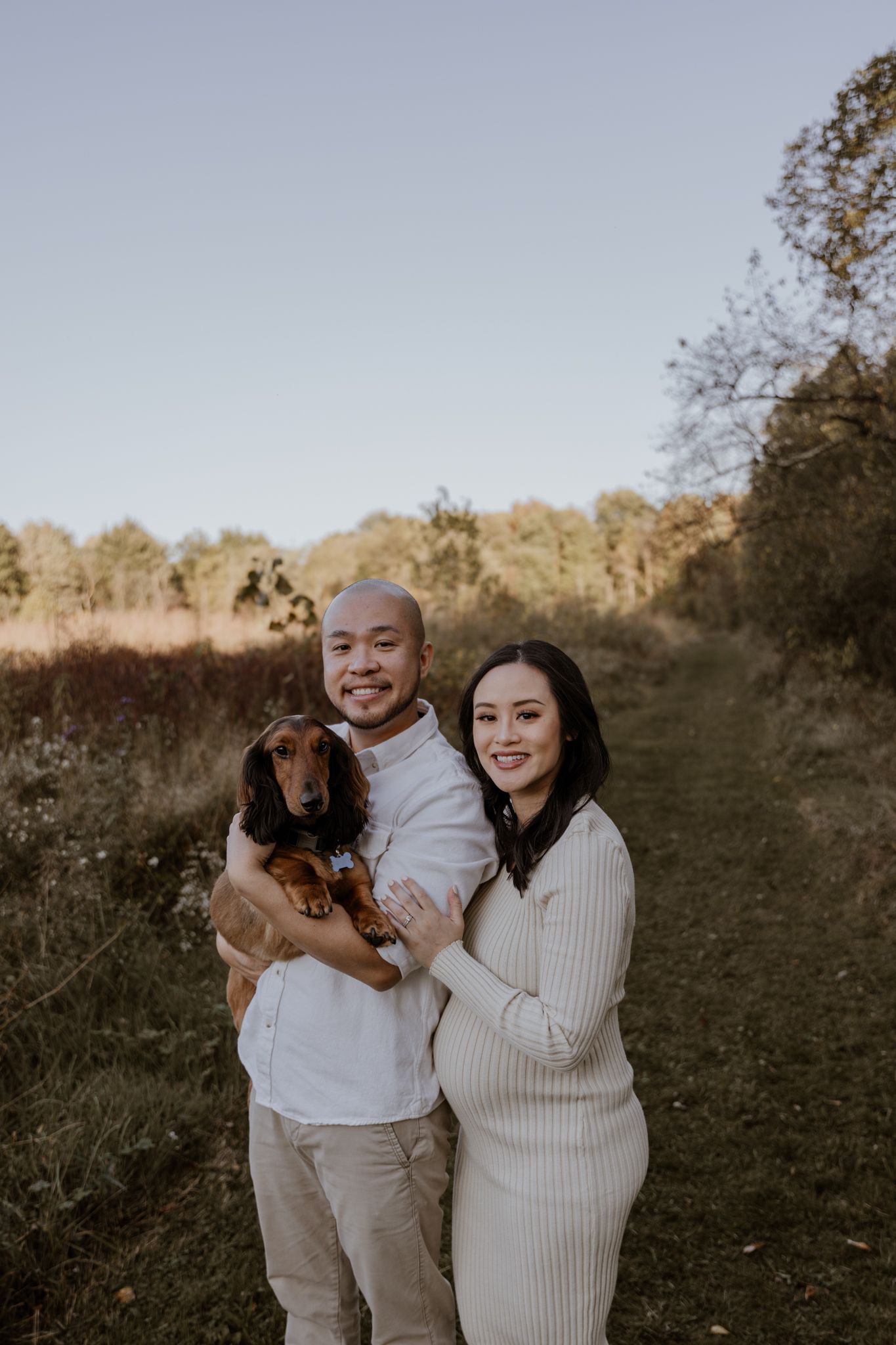 A happy couple poses with their brown dog in a natural autumn setting.
