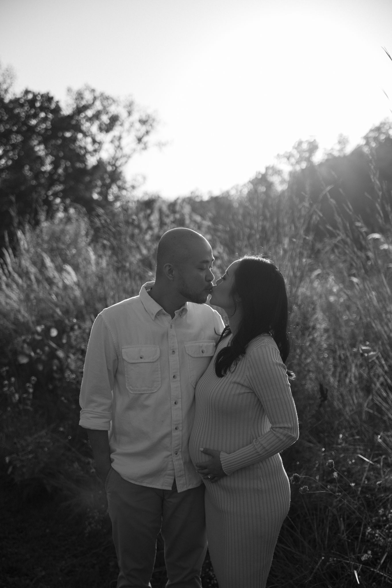 A couple shares an intimate moment in tall grass during a black and white sunset photoshoot.
