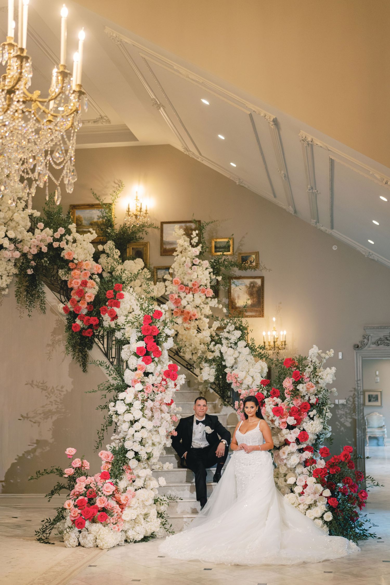 Bride and groom posing on a grand staircase adorned with lush pink and white floral arrangements indoors.