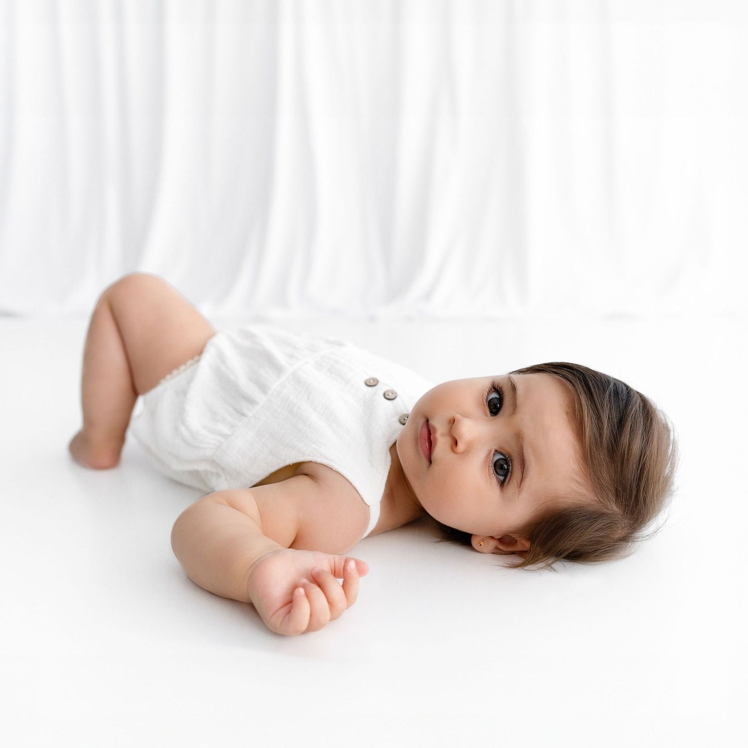Baby wearing white outfit laying on white floor during photo session.
