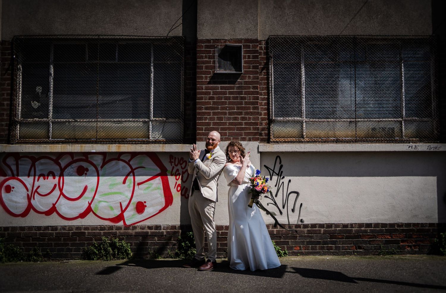 Wedding couple poses against urban backdrop with brick wall and graffiti in casual style.