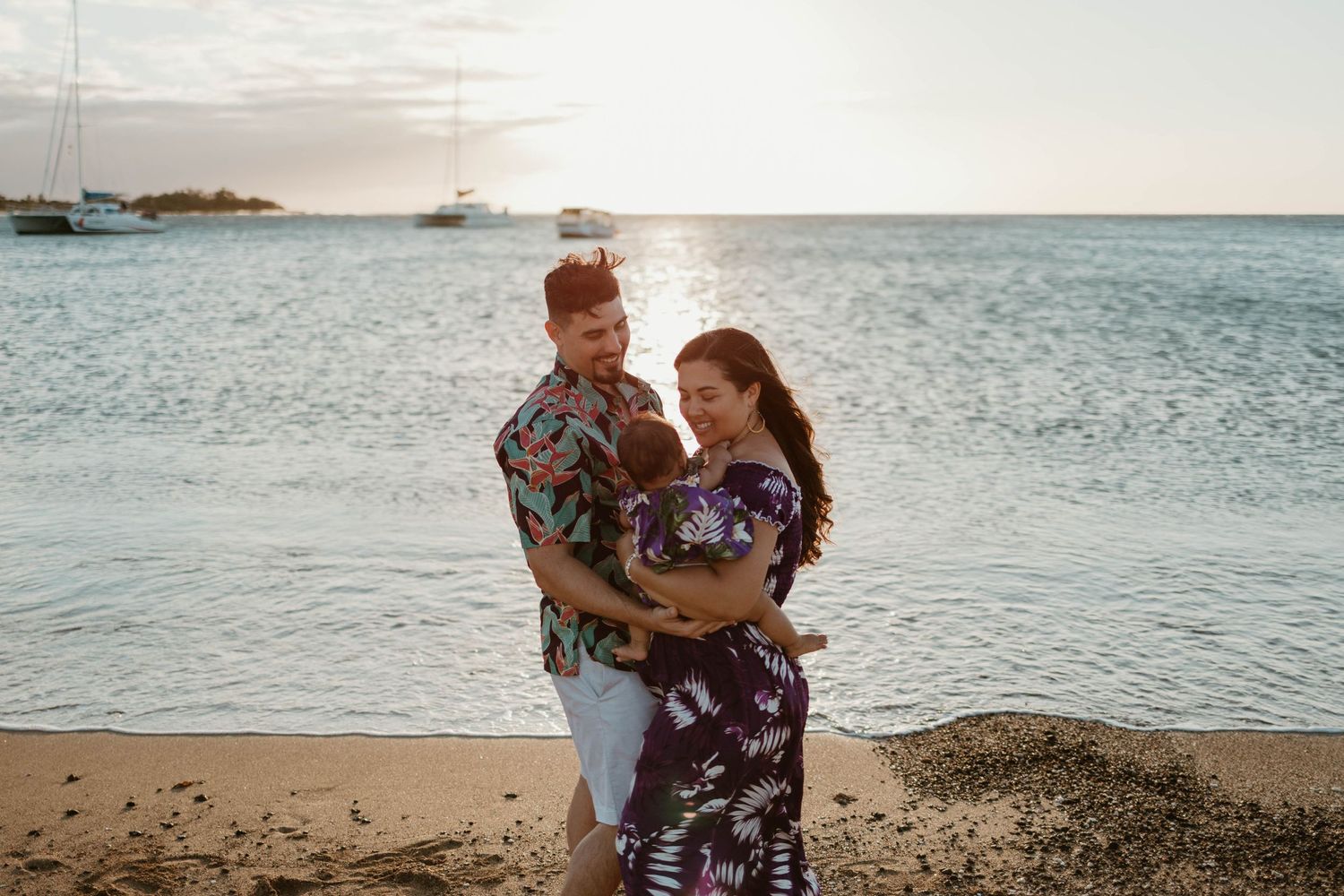 A couple embraces on a beach at sunset with boats visible in the background.