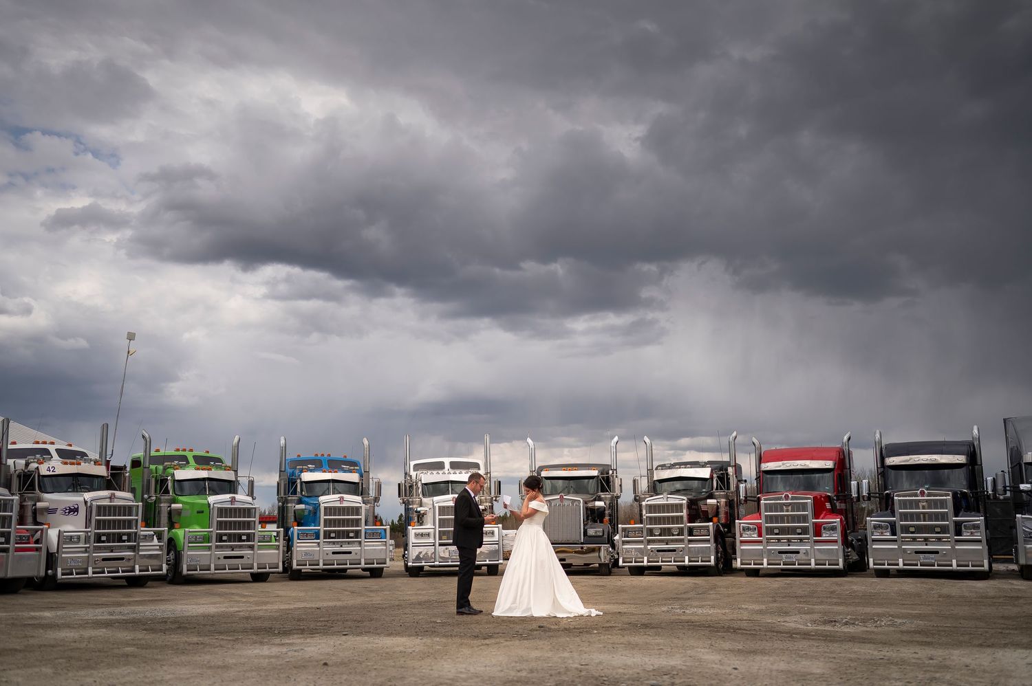Wedding photo with dramatic storm clouds and line of semi trucks in background.
