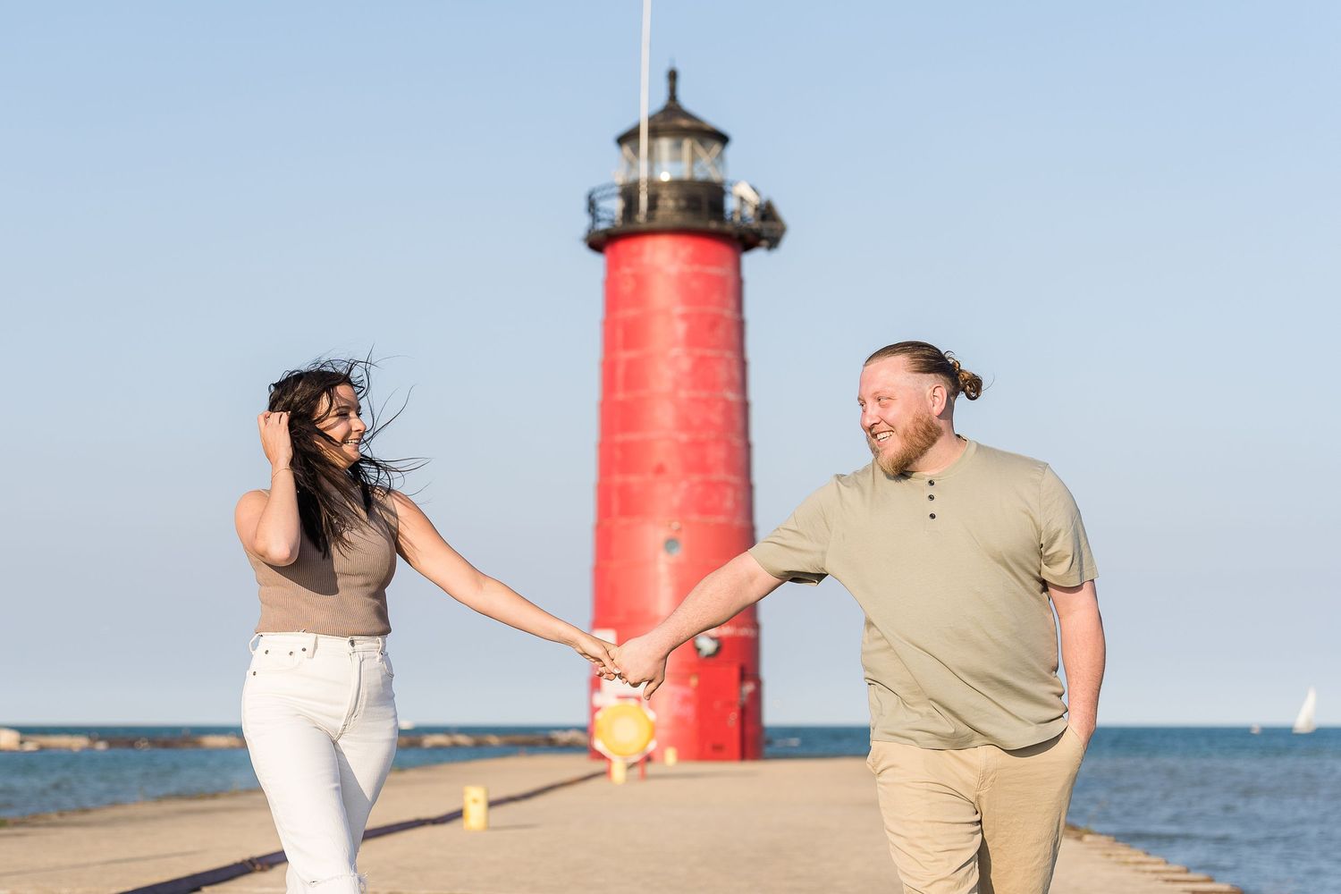 Couple holding hands and laughing in front of red lighthouse on sunny waterfront engagement shoot.