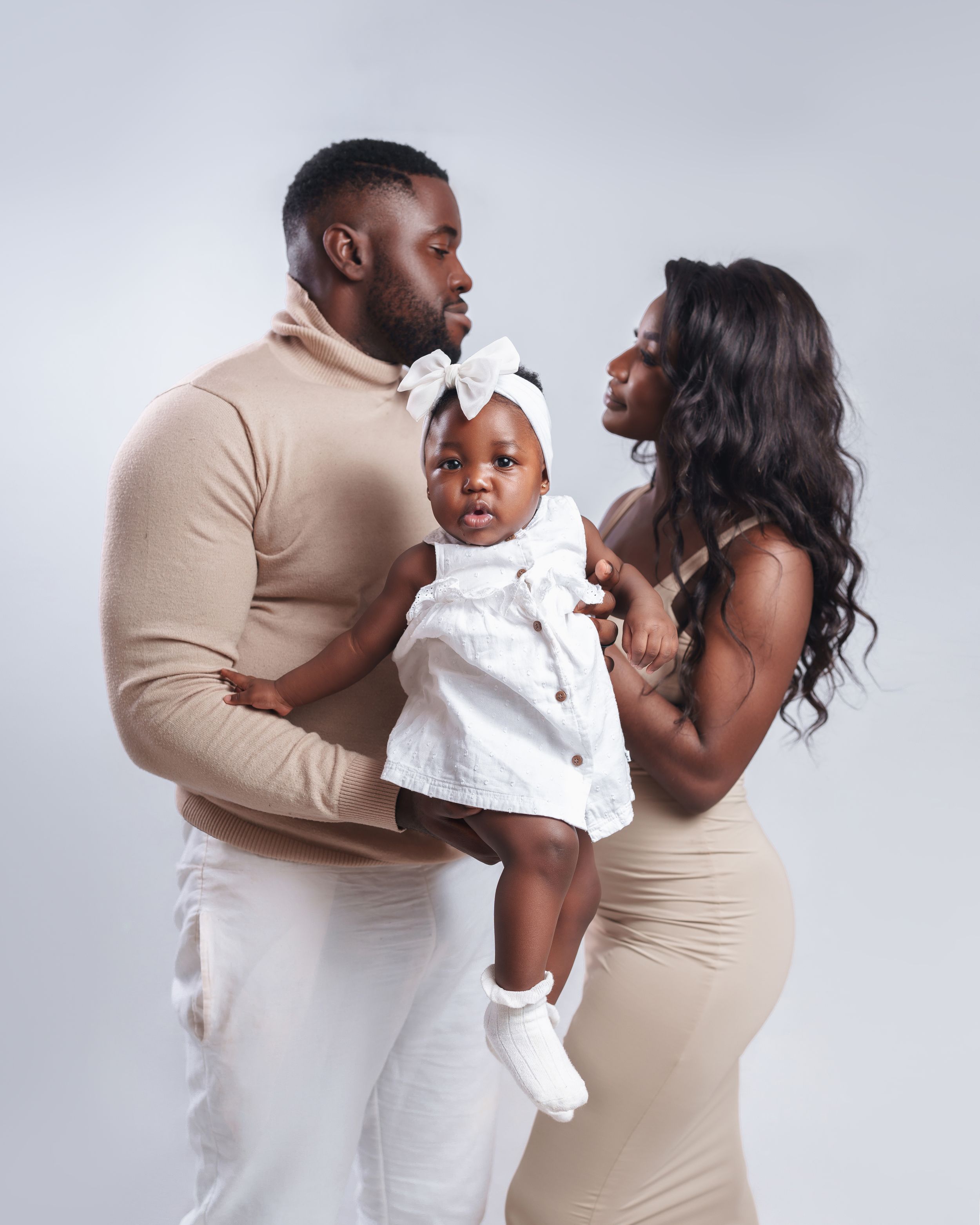 Family of three poses in coordinated beige and white outfits against grey studio backdrop.