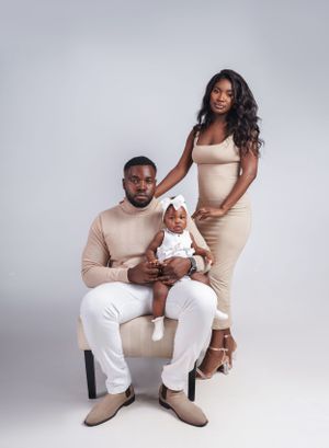 Family portrait features coordinated neutral tones with infant in white dress against grey background.