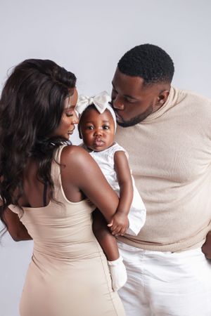 Parents in neutral tones share intimate moment with baby in white dress against studio backdrop.