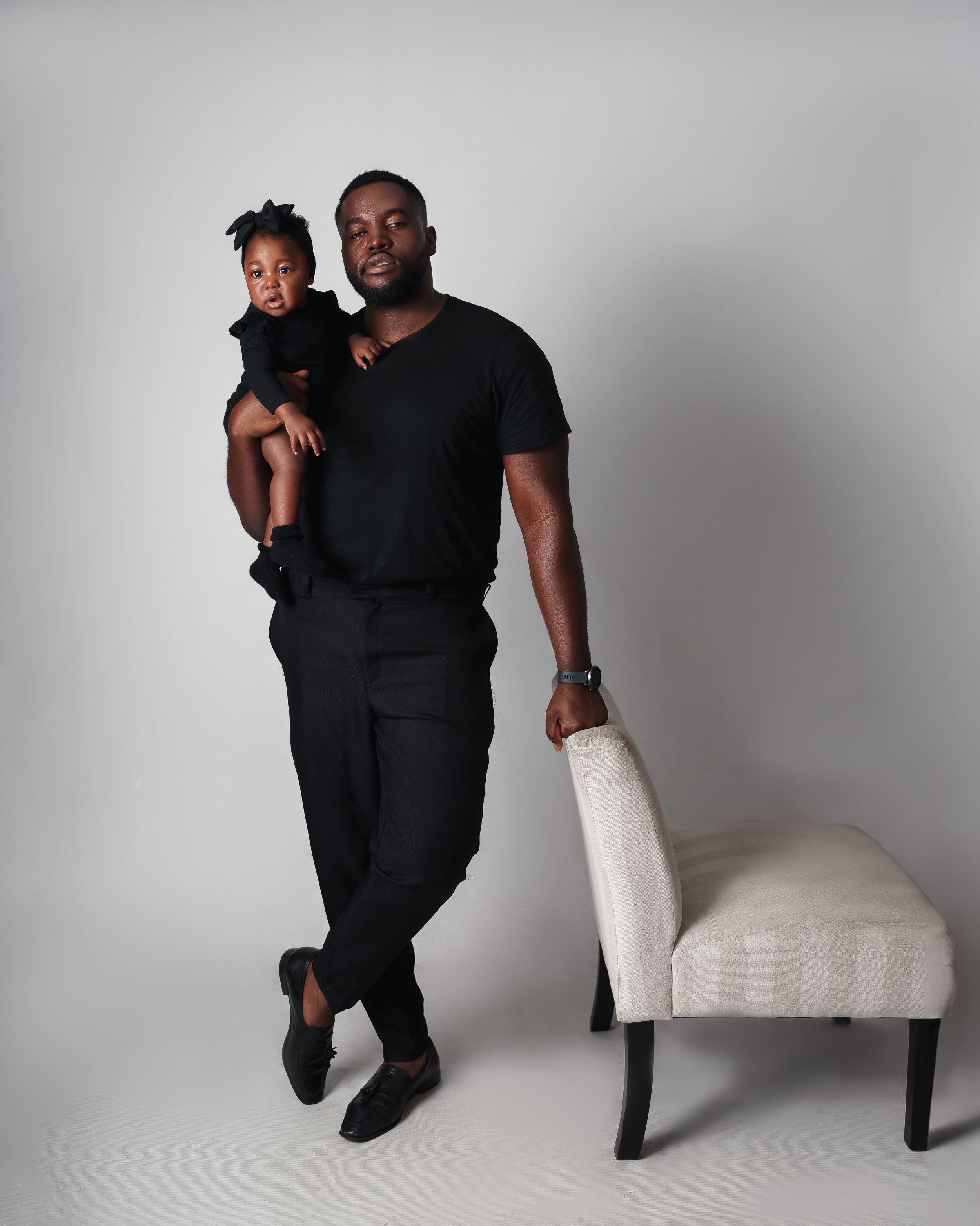 A casual portrait of a parent and child in black outfits next to a white chair.