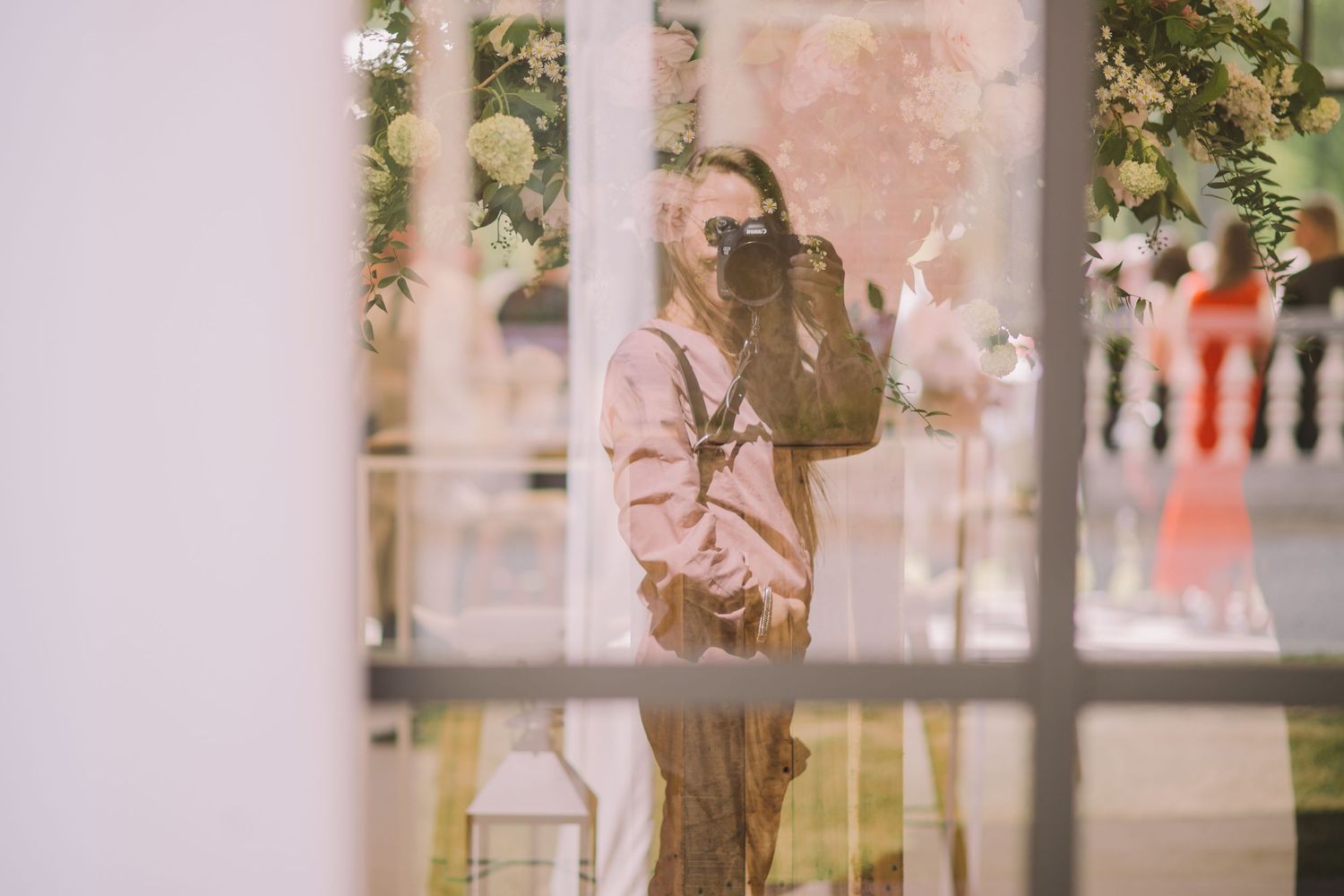 Through a window pane, someone in a pink jacket and mask stands checking their phone in soft natural light.