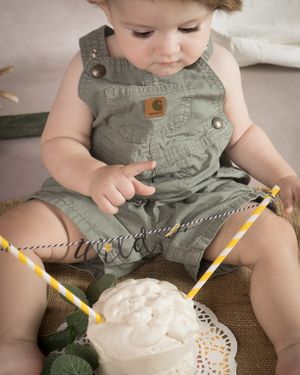 A small child in green overalls sits on a brown surface while reaching for a white cake with a yellow striped straw.