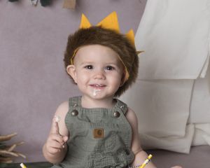 A child in olive green overalls sits on the floor eating cake