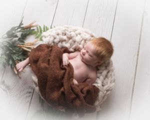 Newborn baby wearing brown teddy bear outfit sleeping on white wooden floor with greenery accents.