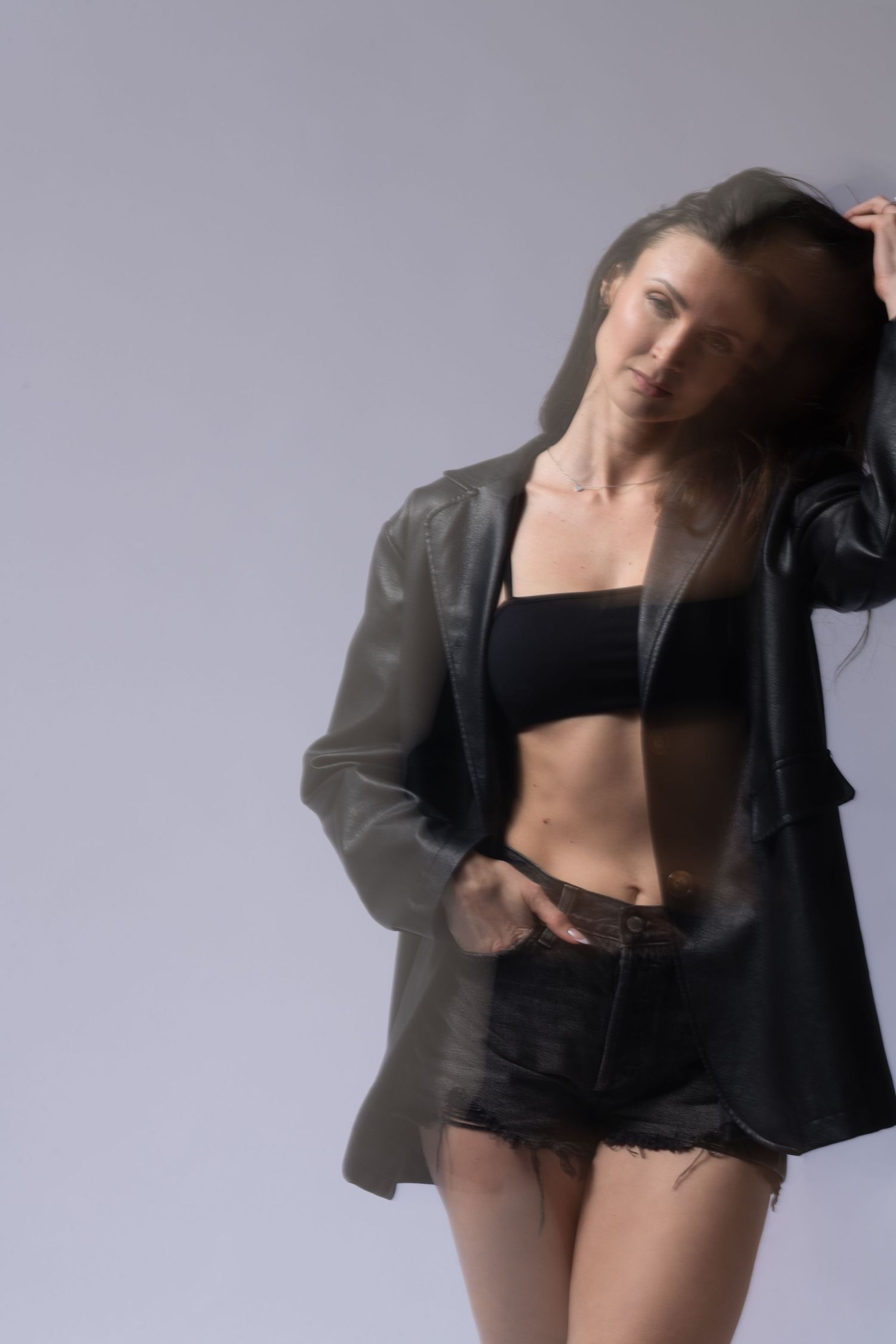 Fashion model in black crop top and denim shorts poses wearing an oversized leather jacket against gray backdrop.