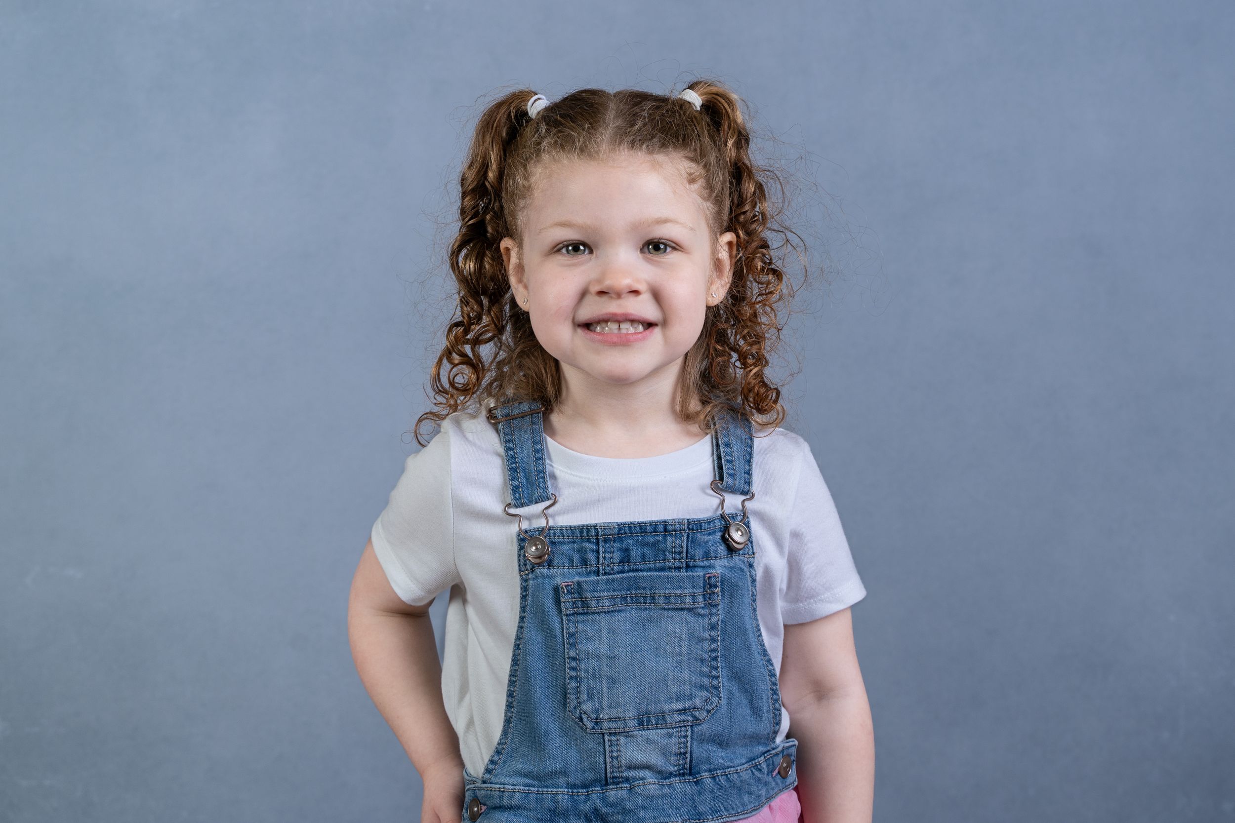 Studio portrait against blue backdrop of young child wearing denim overalls and white shirt.