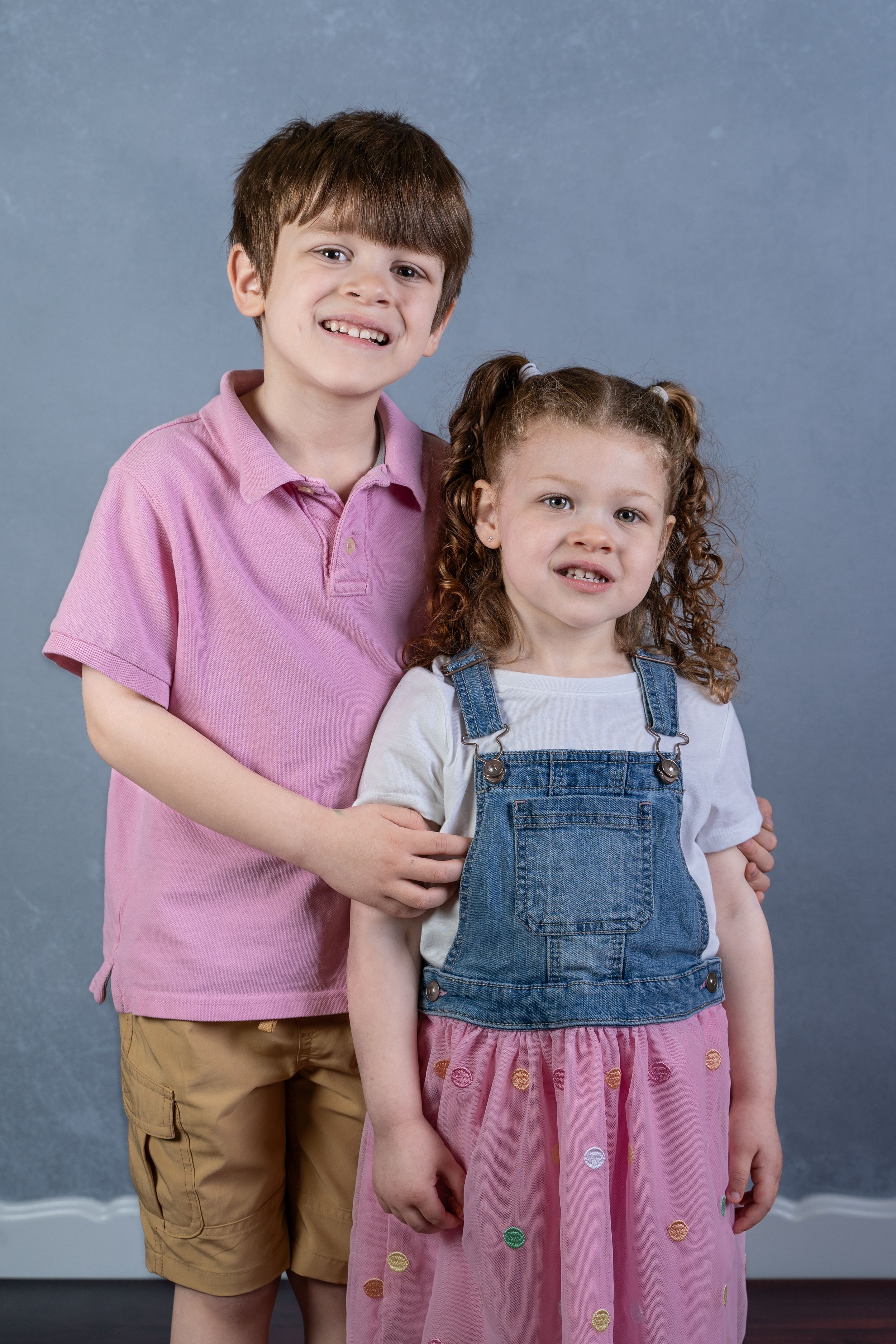 Studio portrait of two children, one in pink polo and one in denim overalls with pink skirt.