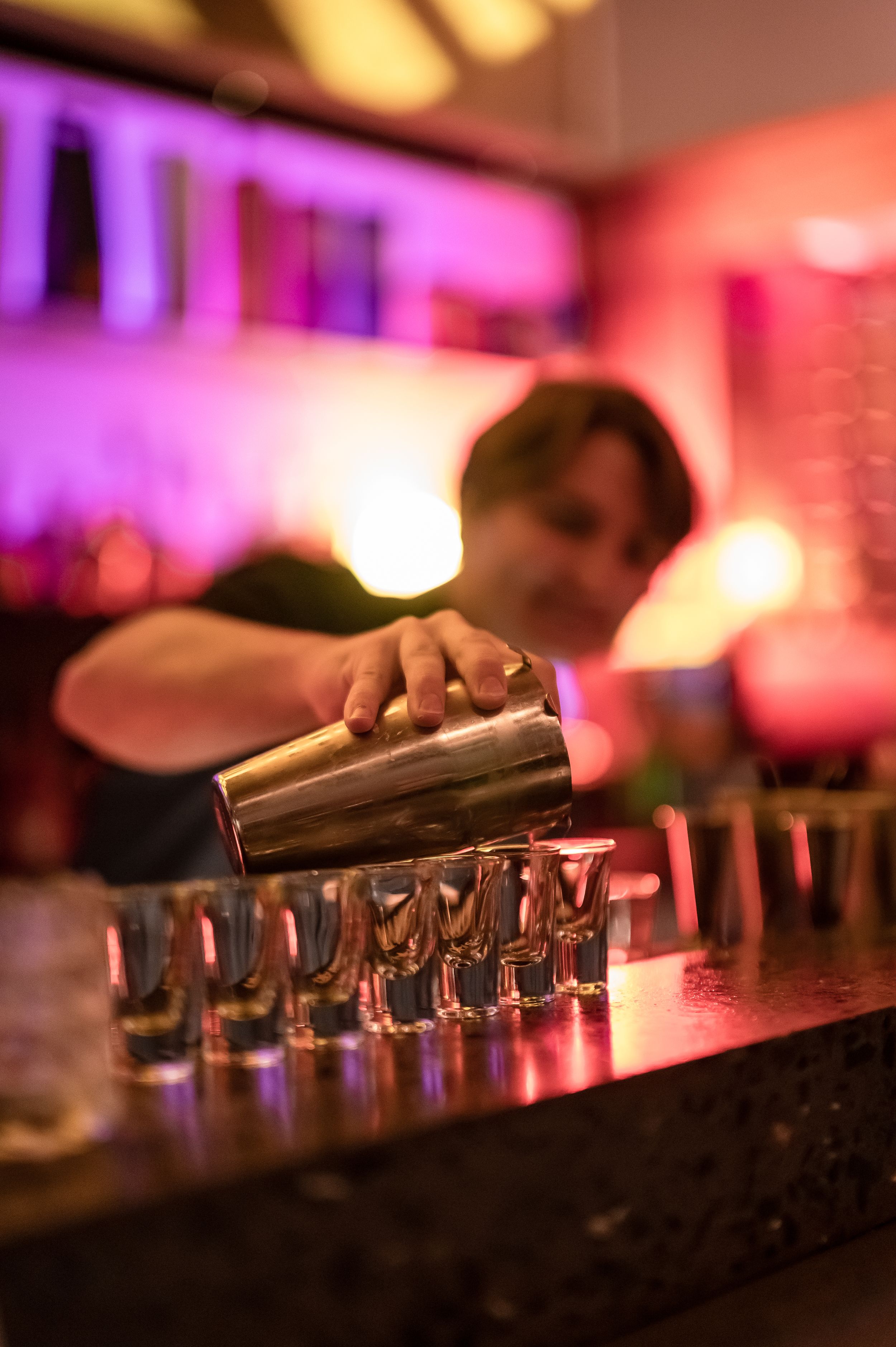 A bartender pours shots from a cocktail shaker into a line of glasses under colorful purple and pink bar lighting.