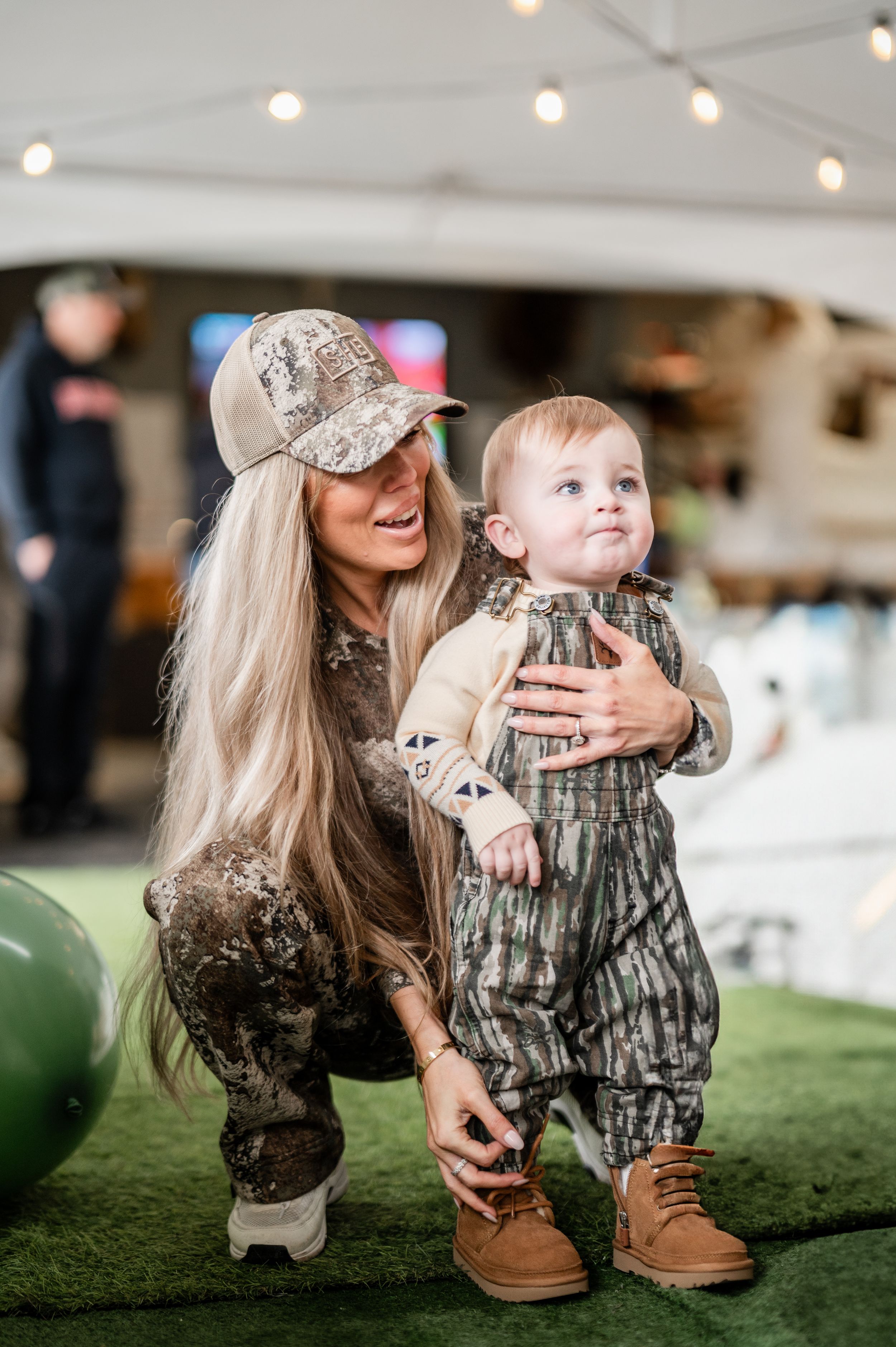 A cute toddler wearing a camouflage outfit and boots stands with an adult in matching camouflage under string lights.