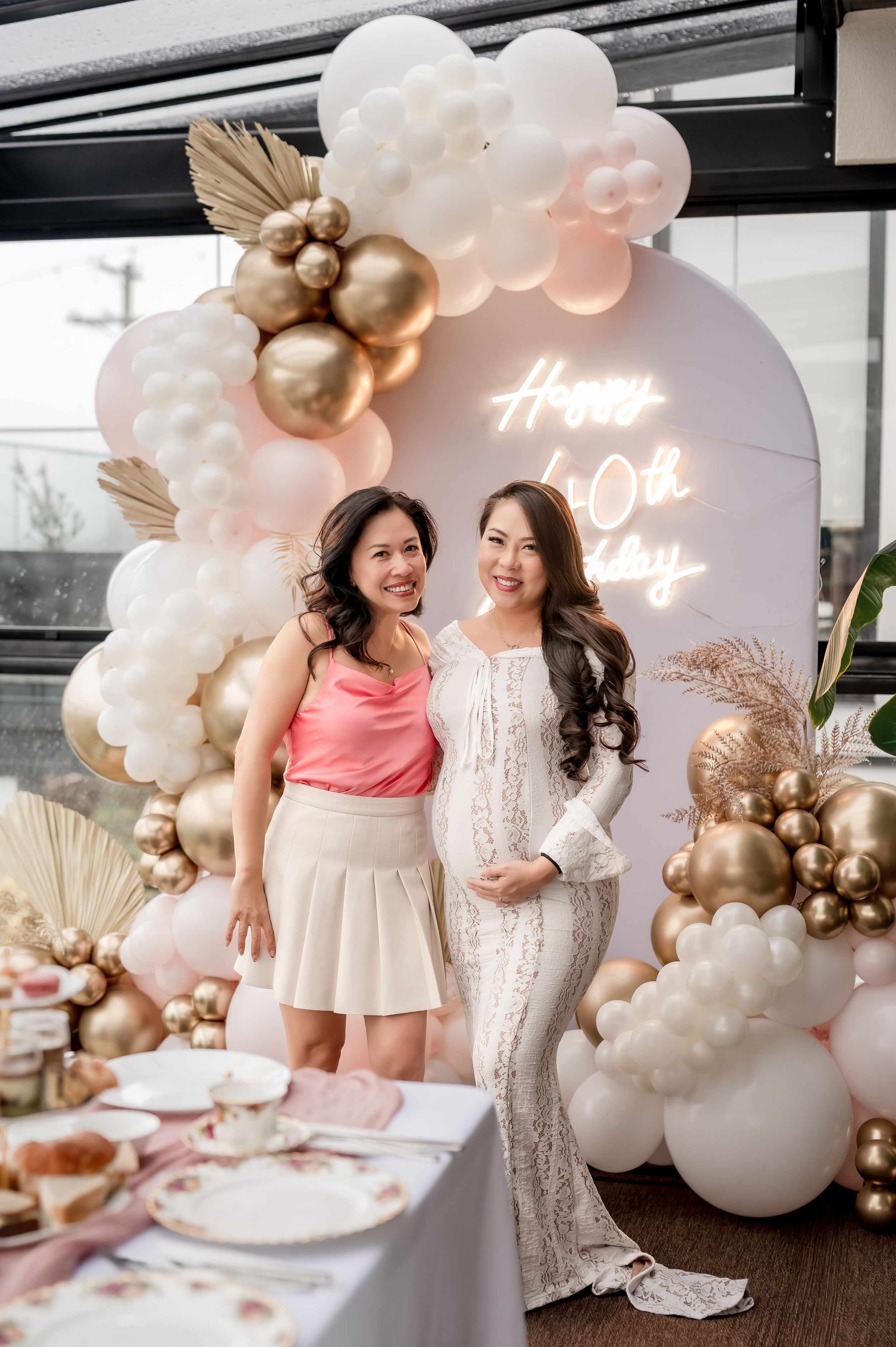 Elegant party setup with pink and gold balloon arch, white lace dresses, and modern decor at a special celebration.