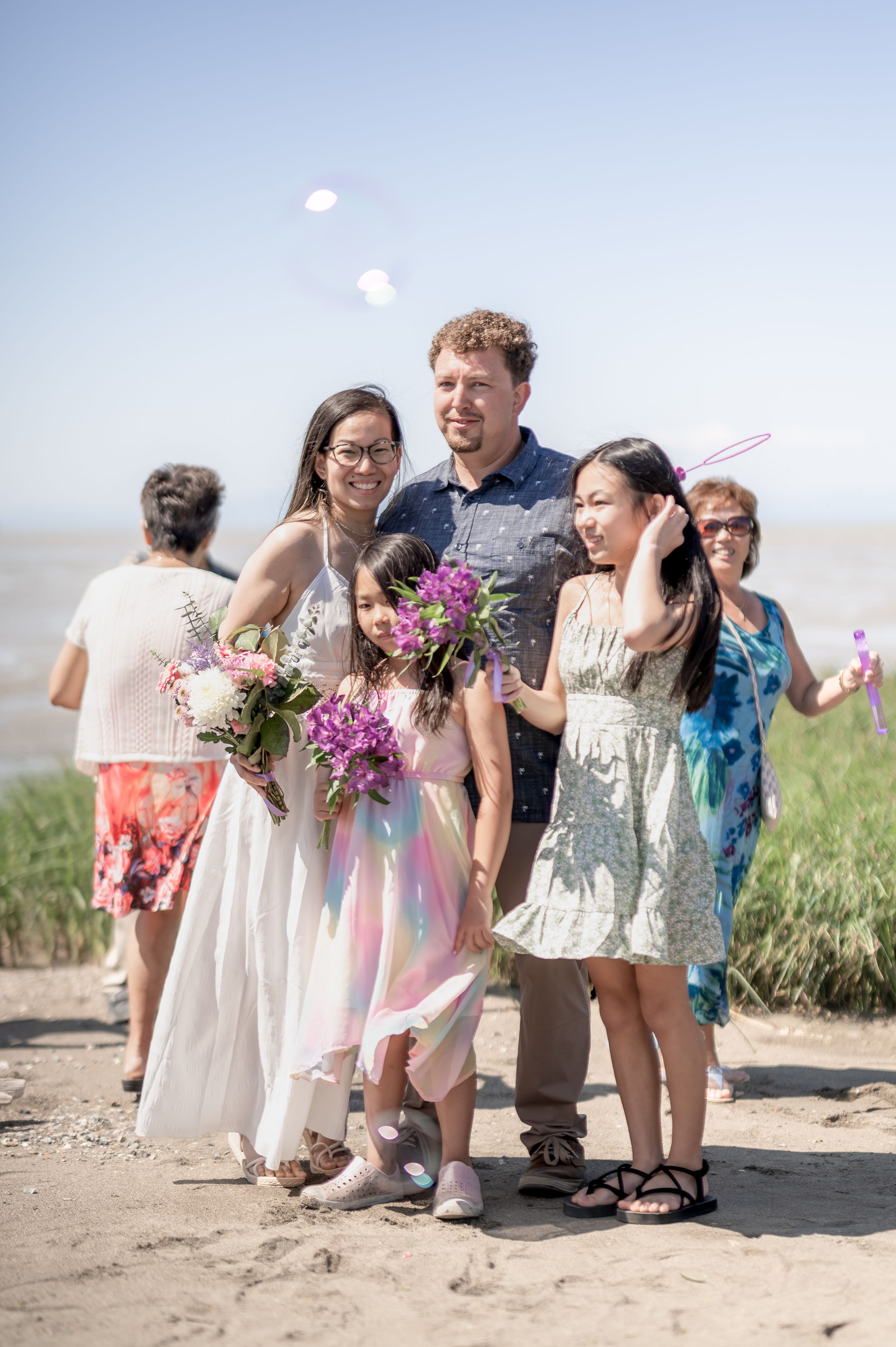 A group enjoys a sunny day outdoors on a beach path, holding vibrant pink and purple flower bouquets.