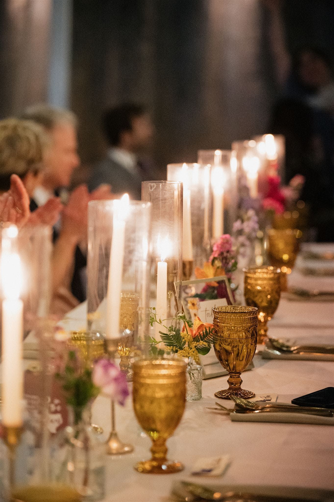Elegant wedding reception table setting with glowing candles, amber goblets, and soft floral arrangements on white linens.