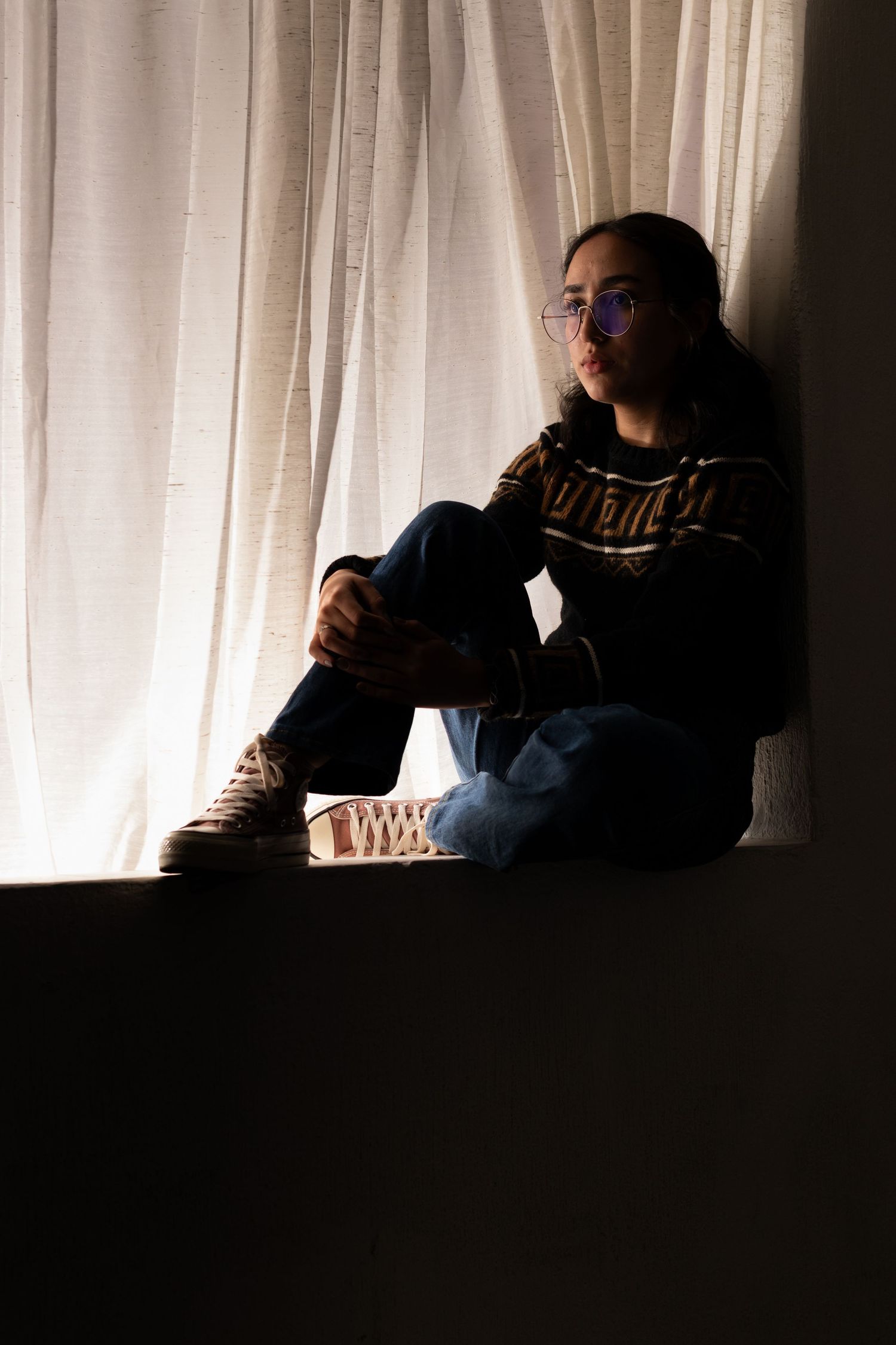 Figure reading by window with white curtains in dramatic lighting.