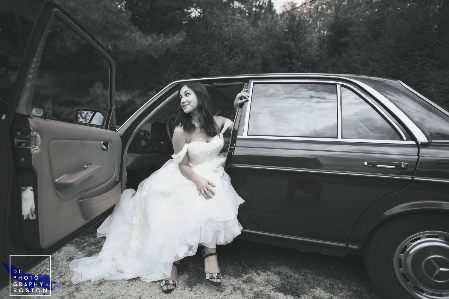 Black and white portrait of a bride in a flowing gown seated in a vintage car.