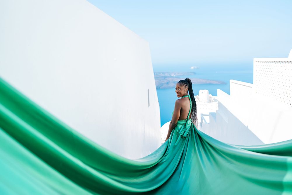 Emerald green dress flows dramatically against white buildings overlooking Mediterranean waters.