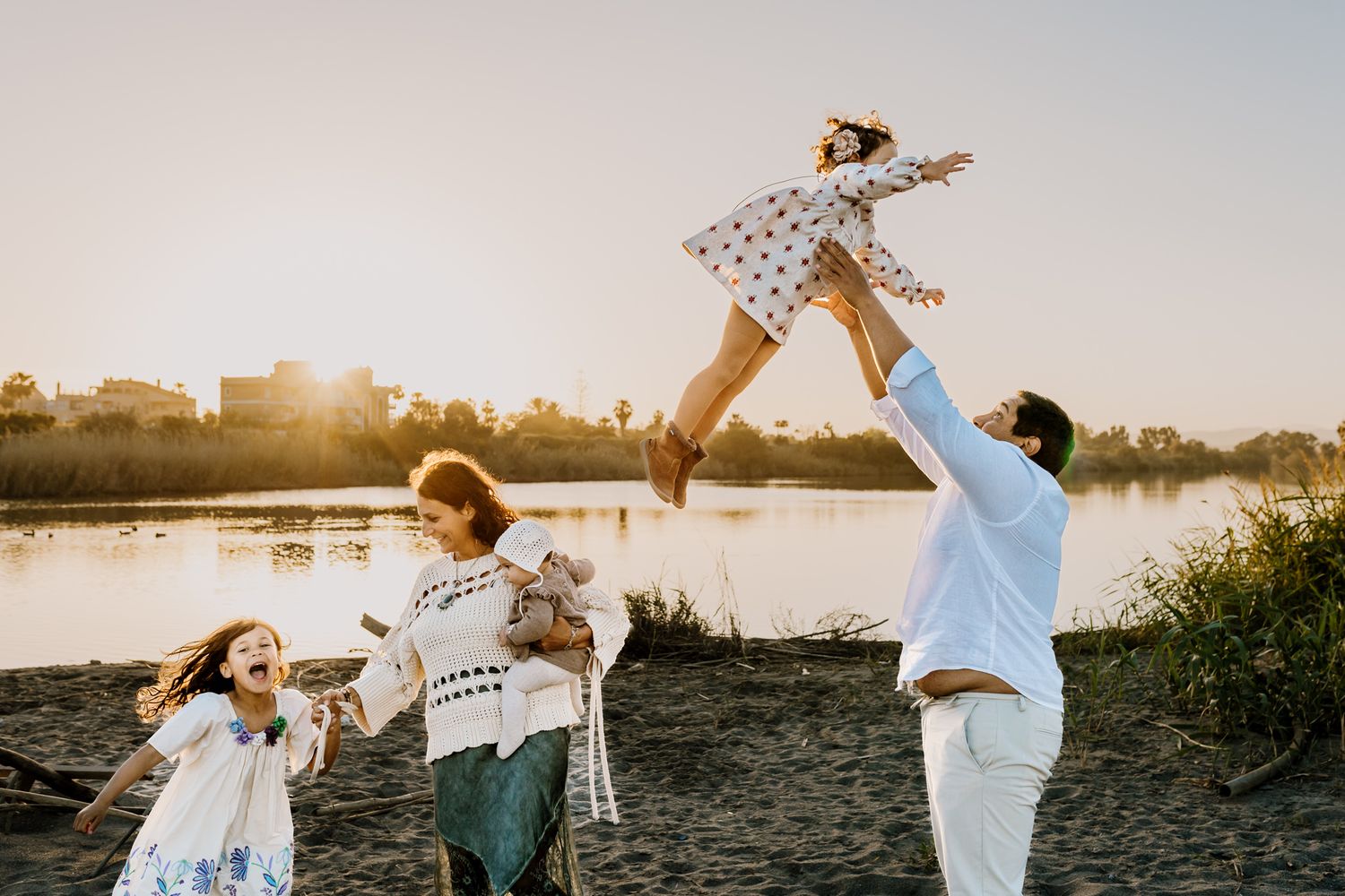Father lift his middle child in the air while mom and her older child and baby are laughing near them.