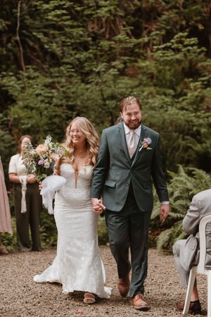 A wedding ceremony takes place on a wooded path with guests seated on white chairs.