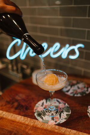 Pouring a citrus cocktail into a coupe glass on a wooden bar surface.